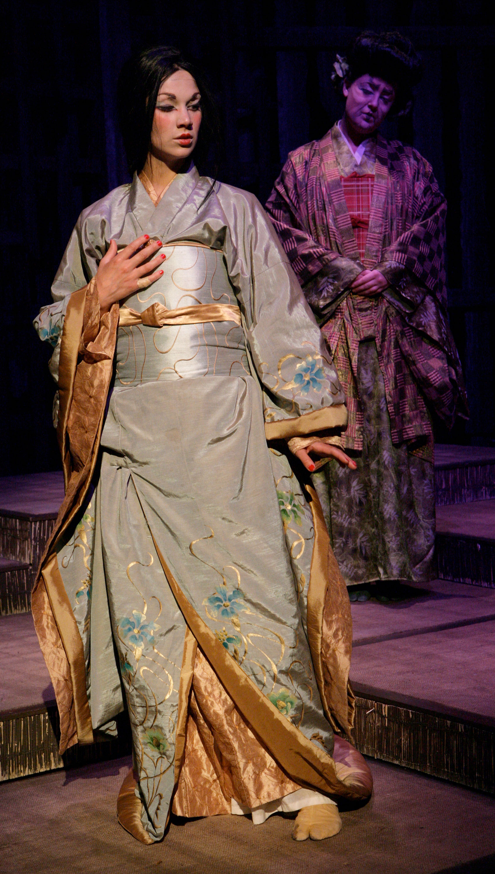 A woman in formal, floral attire accented in gold and makeup is spotlighted on stage. She is looking downward diagonal of stage left and has her right hand on her chest and her left hand at her side with the palm parallel to the floor. An older woman in multi-patterned attire watches the woman behind her from out of the spotlight.