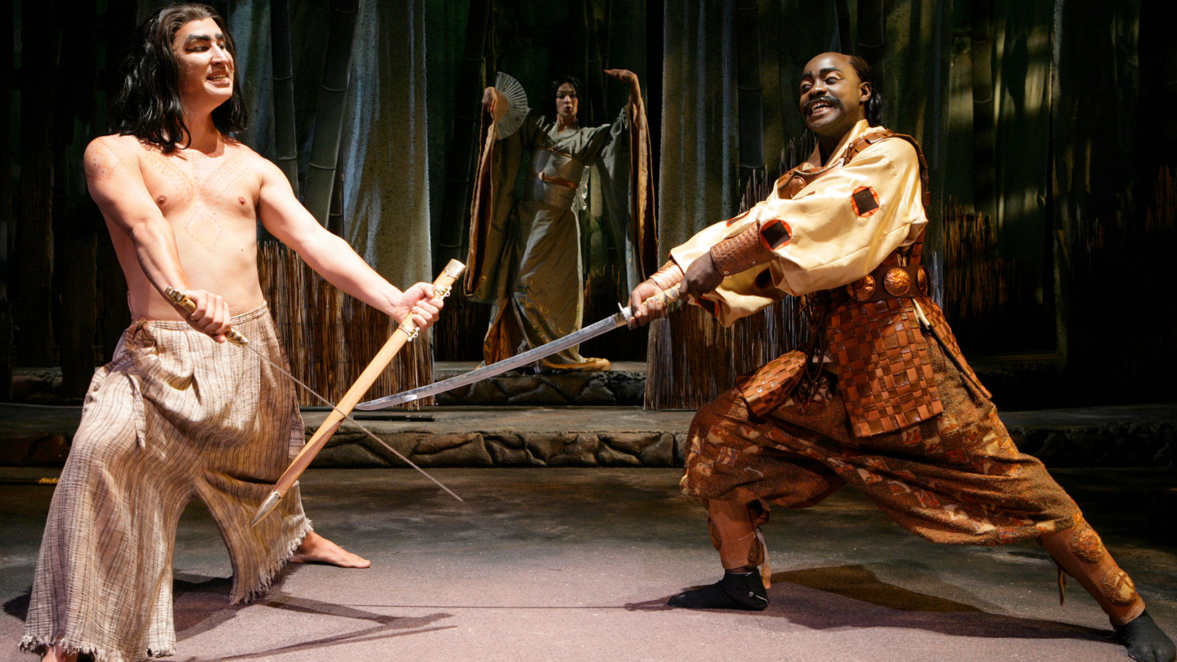 Two men are lunging outward, bodies positioned toward the audience, yet they are looking at each other with wide eyes and bared teeth. The man on the left holds two sharpened wooden sticks with metal points in each hand while the man on the right wields a sword. A woman poses behind them in the background- she wears formal, floral attire with gold accents while holding an open fan held high in her right hand positioned toward her face while she raises her left arm and her palm parallel to the ceiling. She has a look of attitude on her face. 
