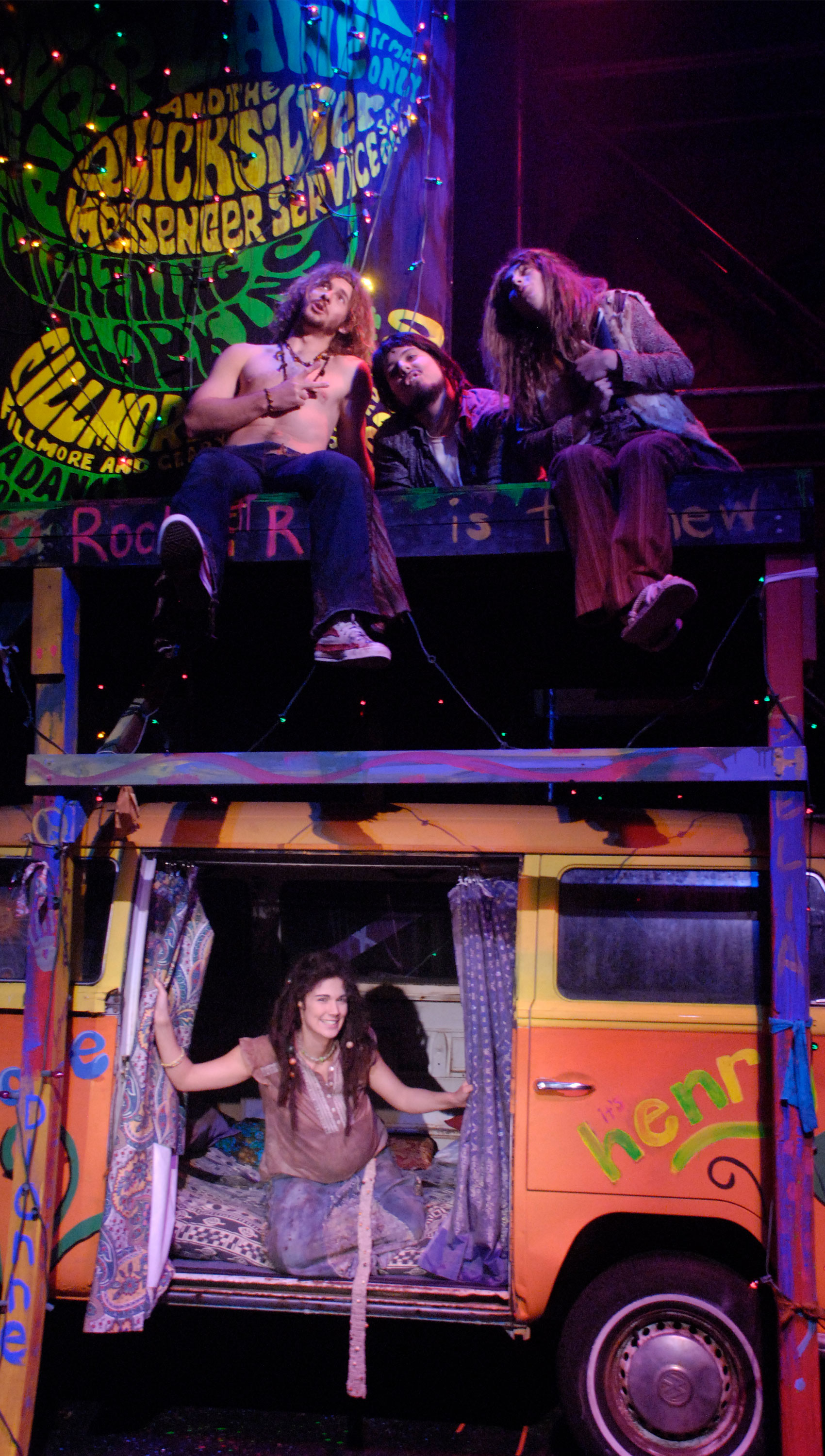 Three characters sit on a platform that is covered in graffiti and are shaping their lips in an “O” shape. They are above a 1960s-style van in which the side is opened and curtains are being pushed aside by a pregnant woman smiling at the camera.
