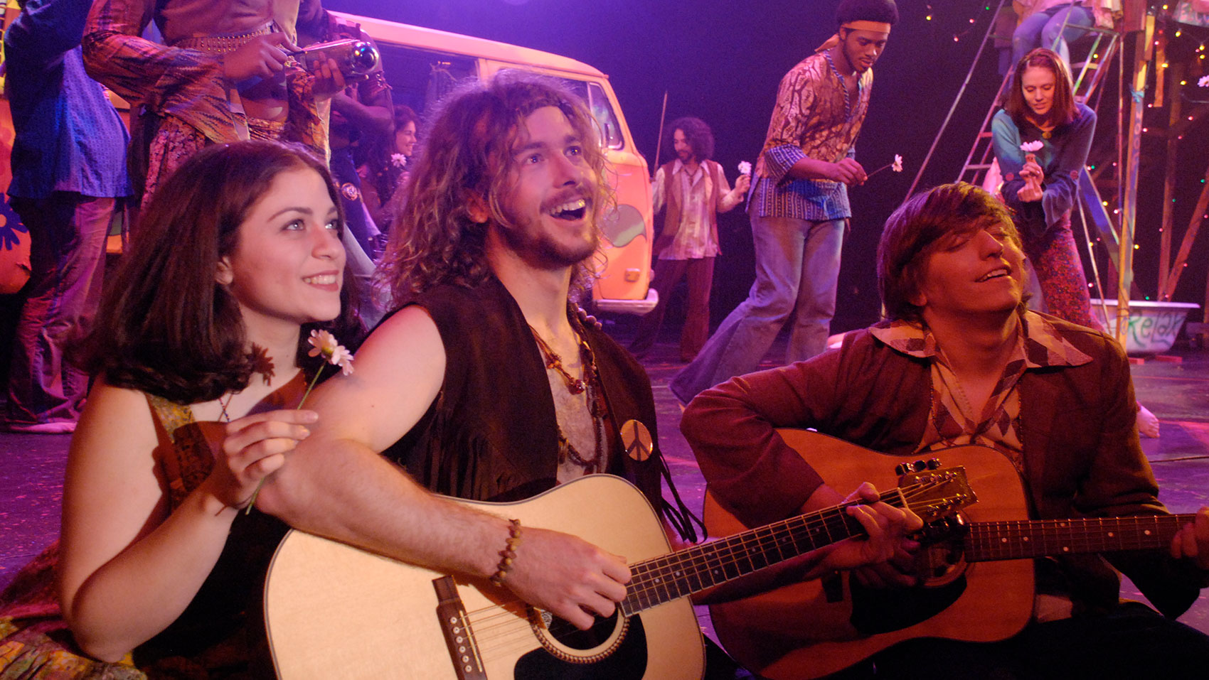 Two men sing with a smile while playing a guitar, one with his eyes shut and the other sitting straight up while looking off into the distance, while a woman sits to the left of the upright man, smiling and looking off into the same direction as the man and holding a daisy. 