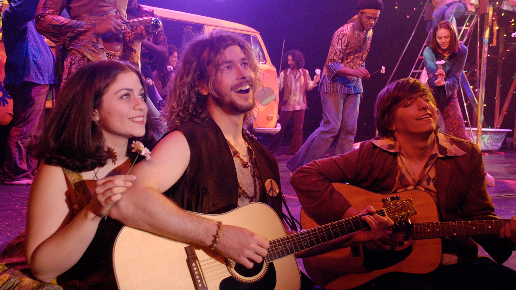Two men sing with a smile while playing a guitar, one with his eyes shut and the other sitting straight up while looking off into the distance, while a woman sits to the left of the upright man, smiling and looking off into the same direction as the man and holding a daisy.