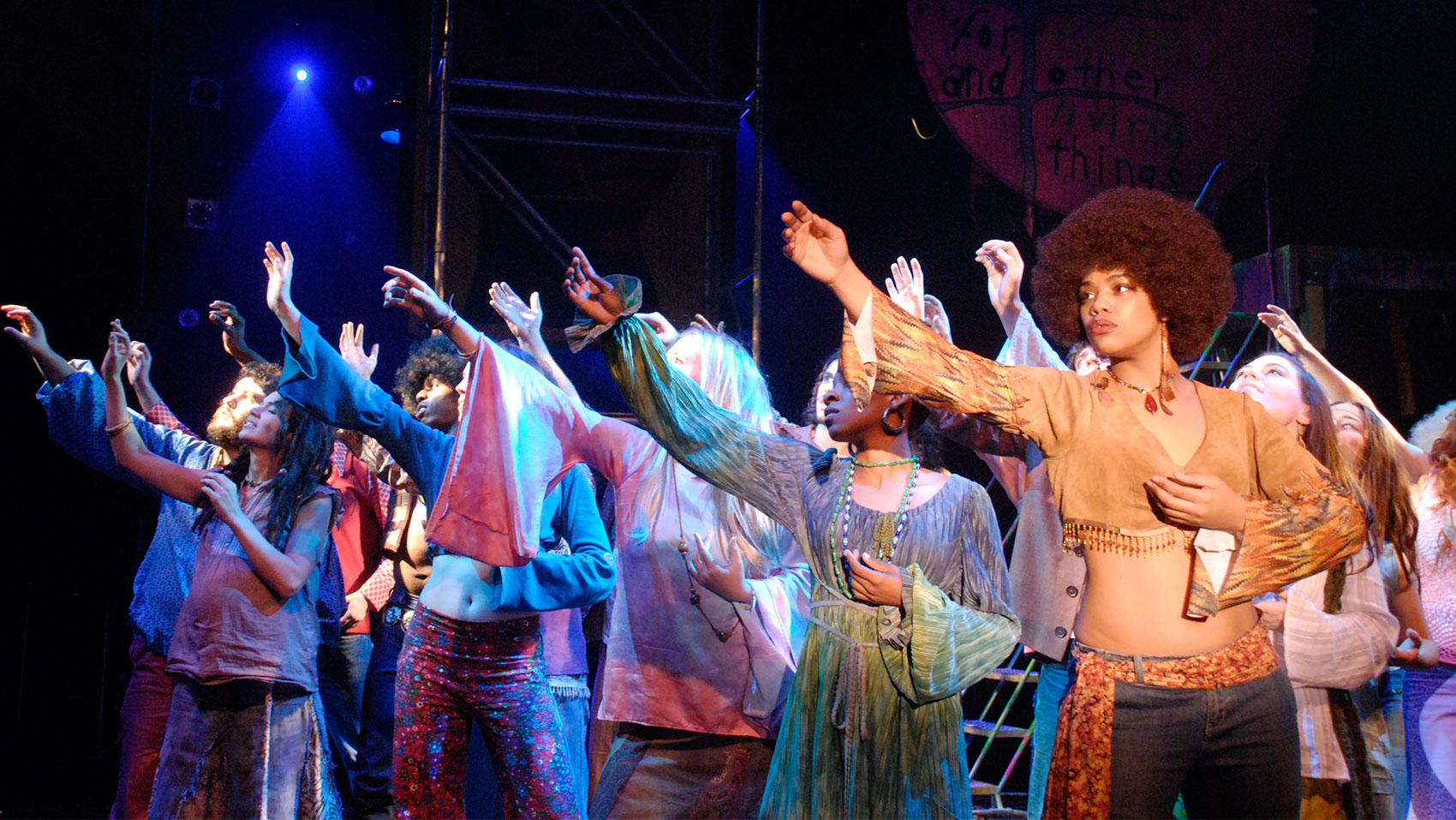 A group of characters in brightly colored costumes stand onstage looking out toward the audience and holding their right arm outstretched as if reaching for something. 