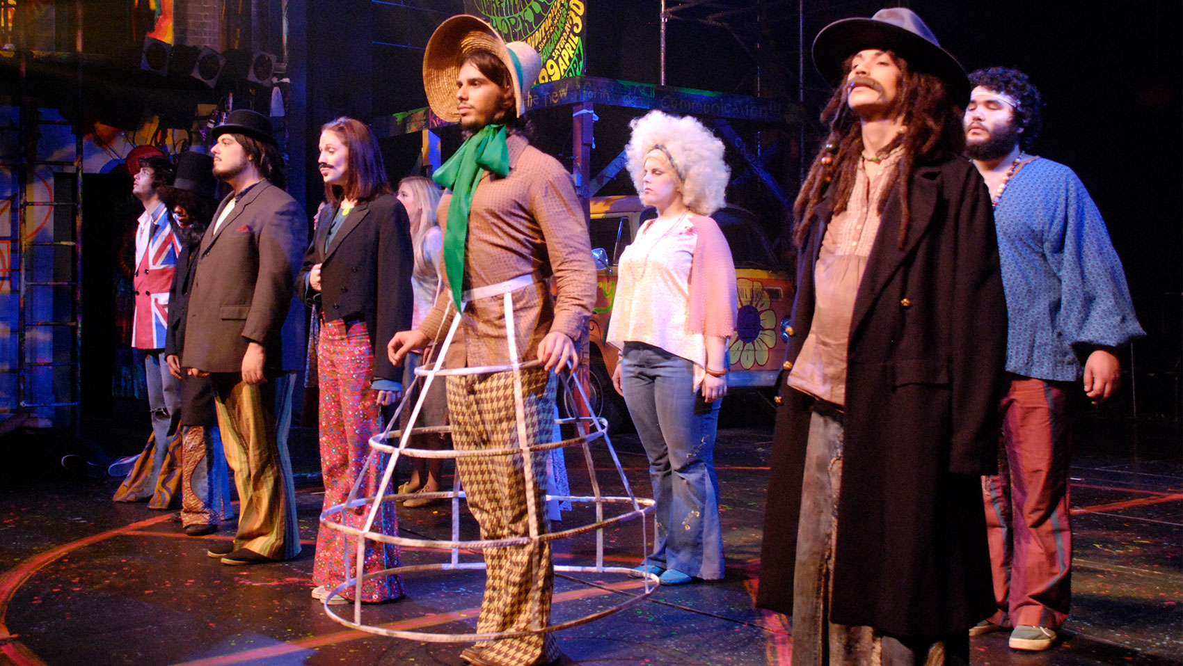 A series of eccentrically costumed characters stand in two lines onstage.