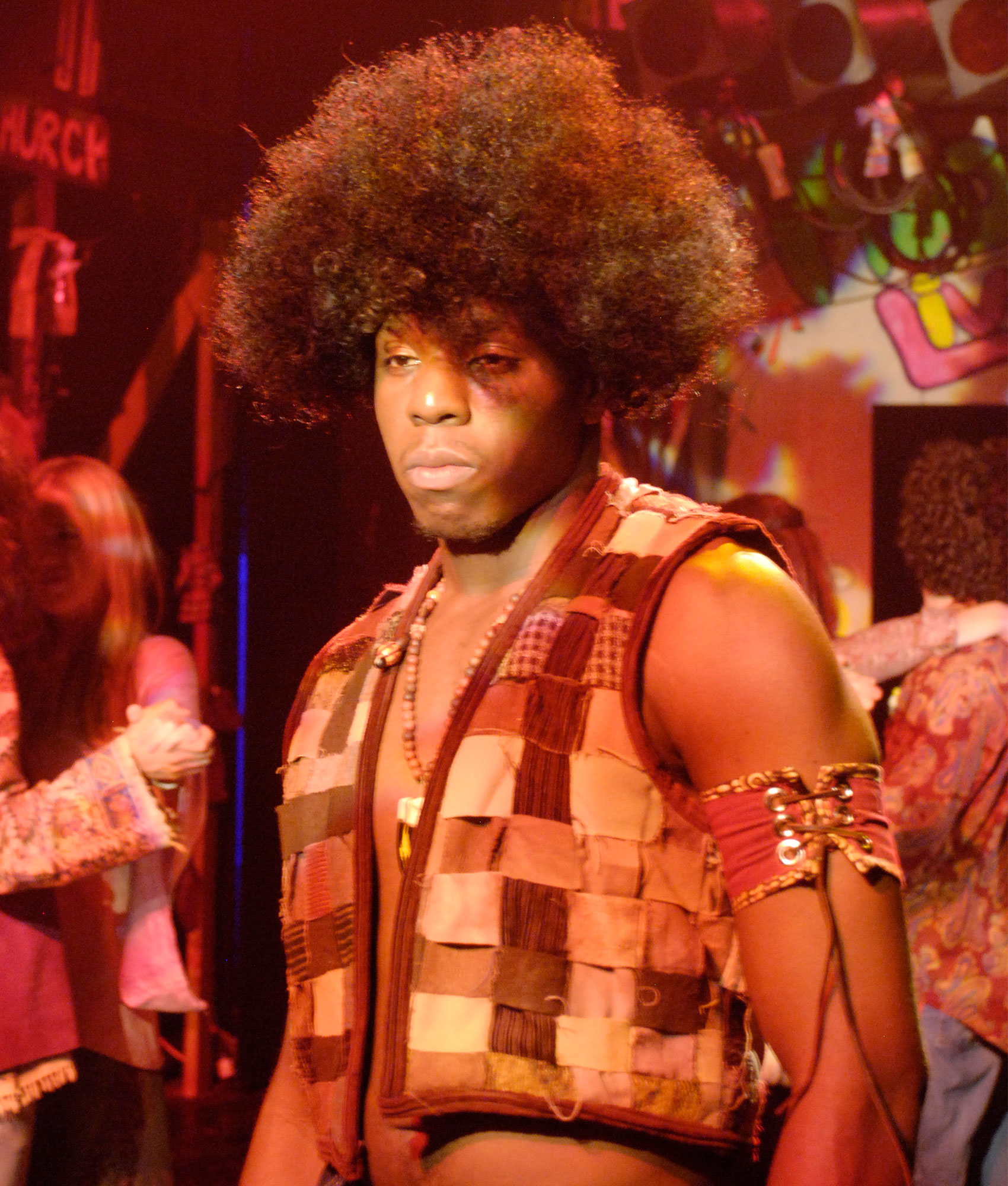 A close-up of a man with a solemn expression and an afro wearing a patchwork vest and a laced-up band around one of his triceps.