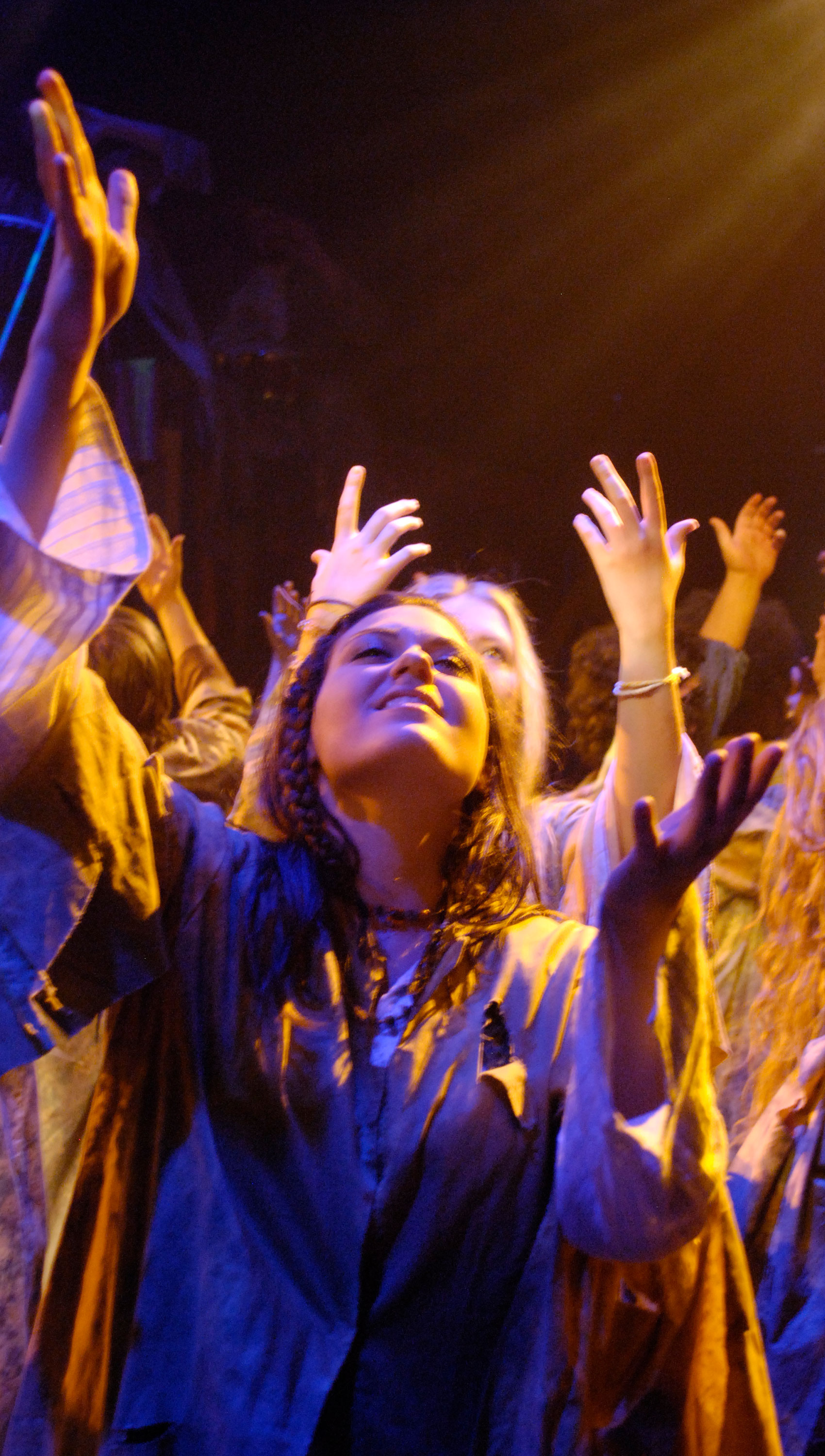 Characters are grouped together on stage raising their hands reverently. A woman looks up with reverence on her face. 