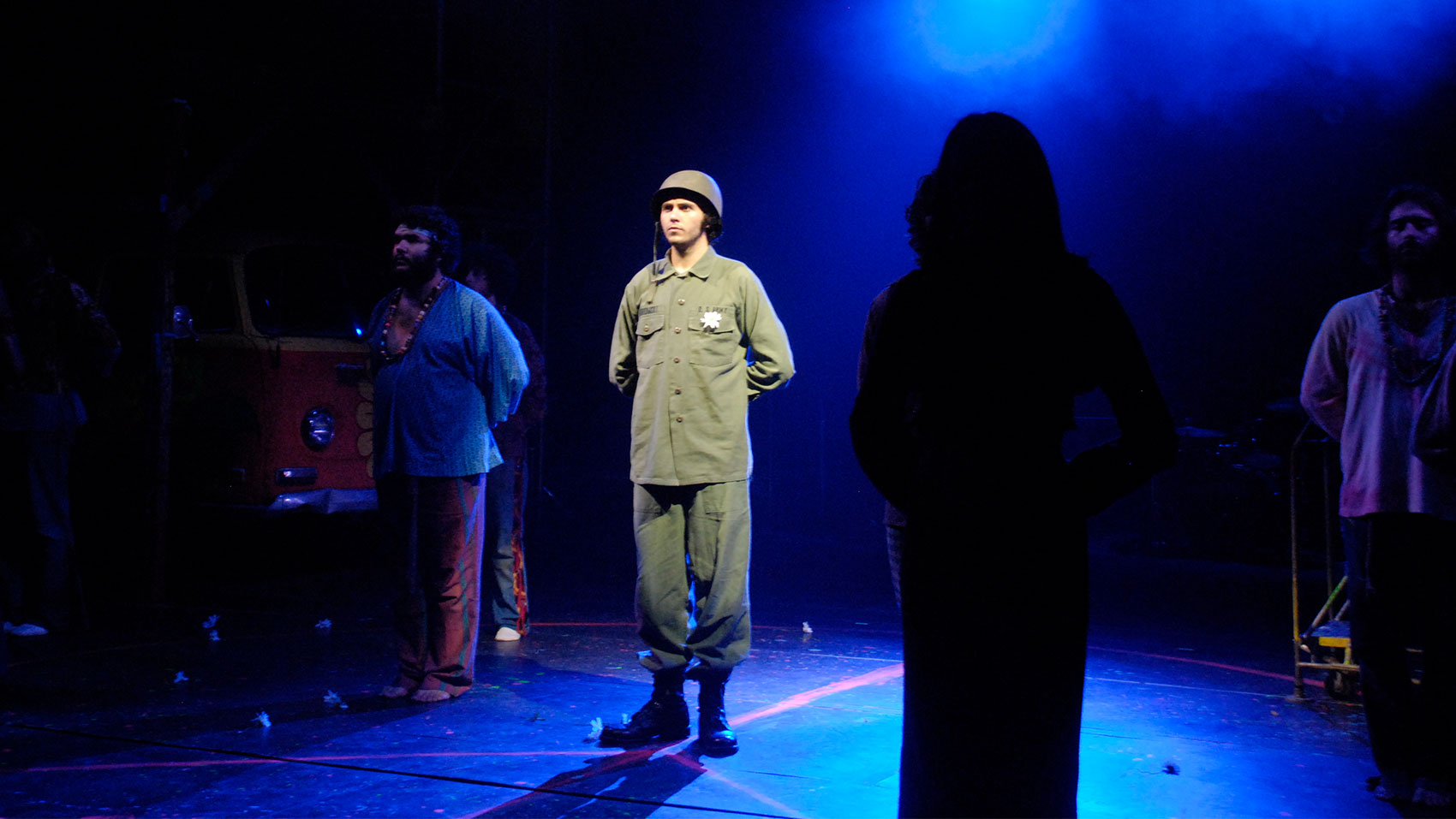 A group of characters are standing on stage, the character furthest into the foreground is silhouetted while a soldier in the mid-ground is under a bright spotlight.