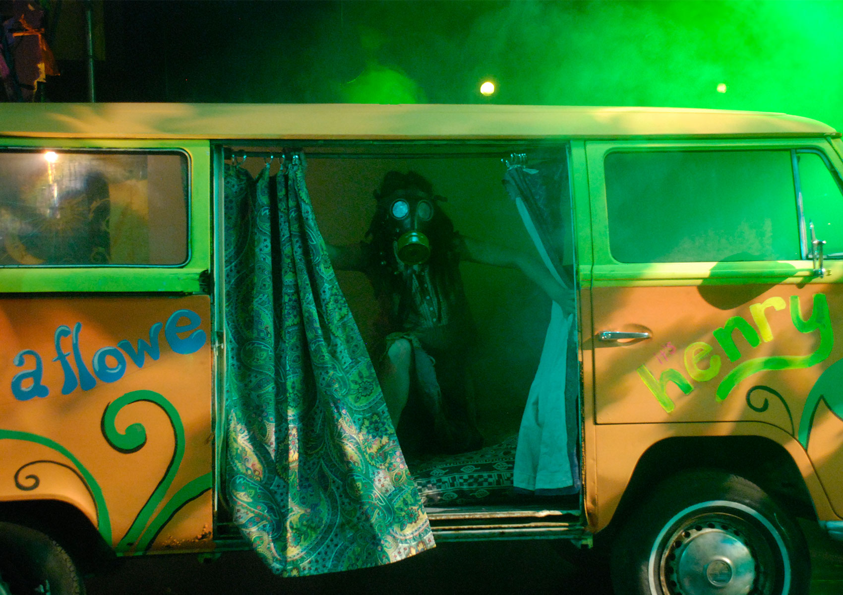 A woman pushes aside the curtains on the side of a 1960s van while wearing a gas mask and being spotlighted by green light.