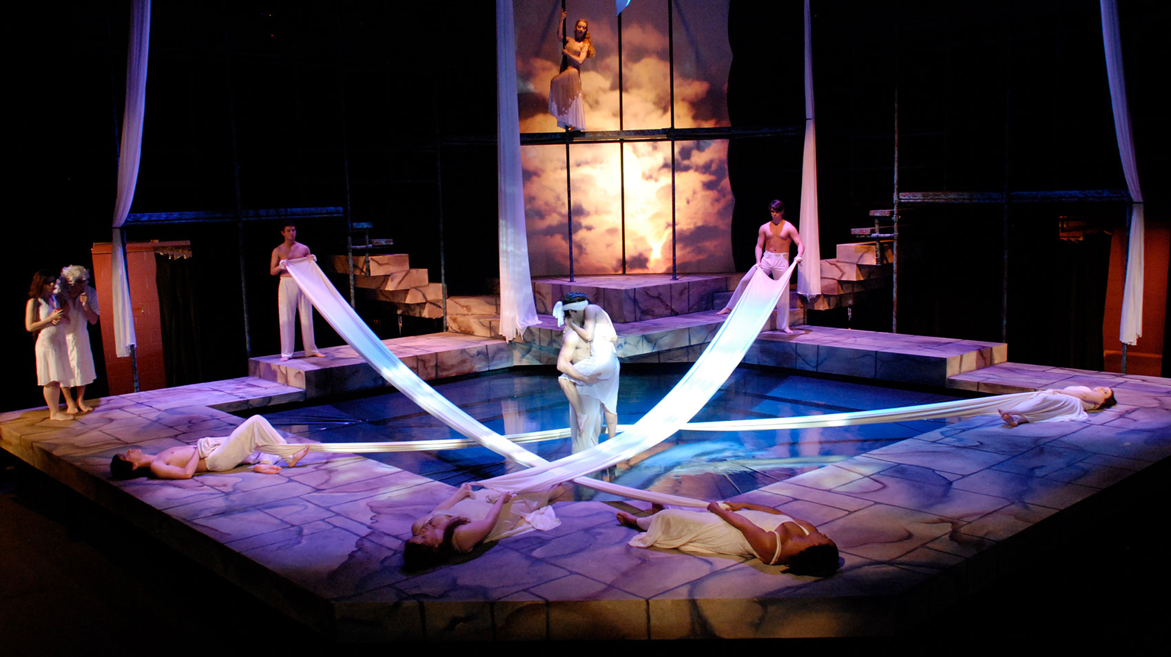: A blindfolded man presses a woman to him in n embrace by supporting her underneath her leg and her back while she has her arm wrapped around his neck and her head behind his. Characters in white attire surround the couple, six surrounding them by holding white drapes stretched end-to-end across a pool in the middle of the stage. Two women in white watch from the background while one watches from a platform above stage.