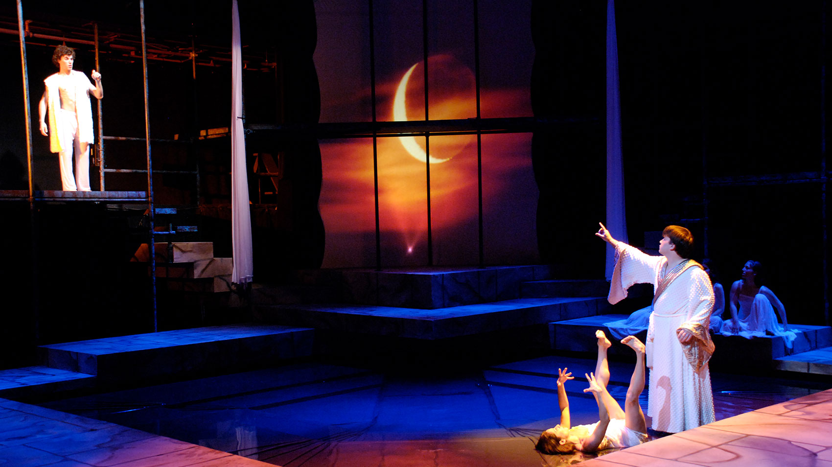 Two characters are standing on the edge of a pool in the middle of the stage, one lying on their back with their arms and legs in the air and the other standing over the first, looking up and left while pointing at a third character standing on a platform looking down at the second character with his right hand and index finger pointed in the air.