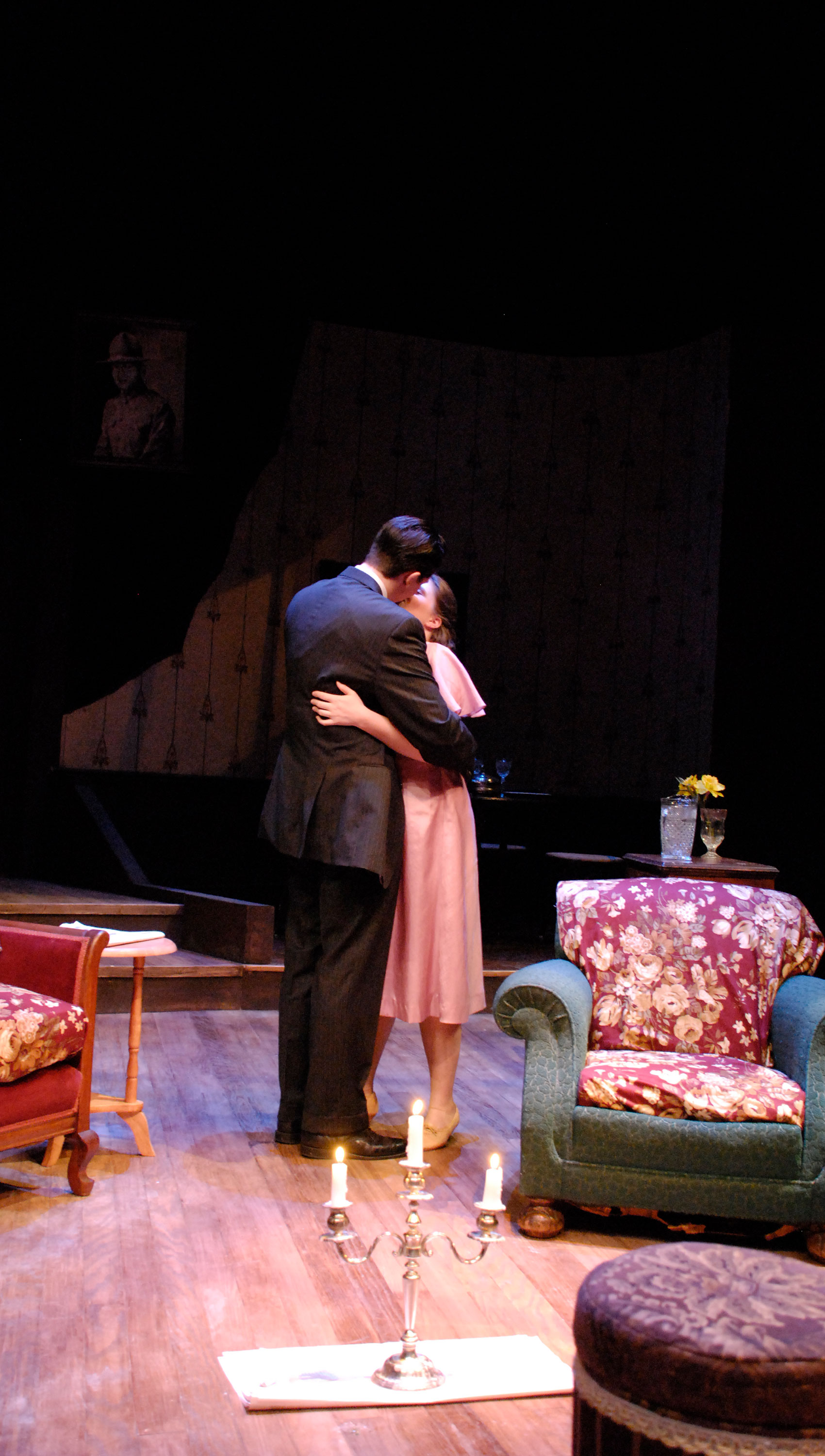 A young man and woman hold each other as they kiss standing in a living room, a lit candelabra on the floor in front of them upstage.