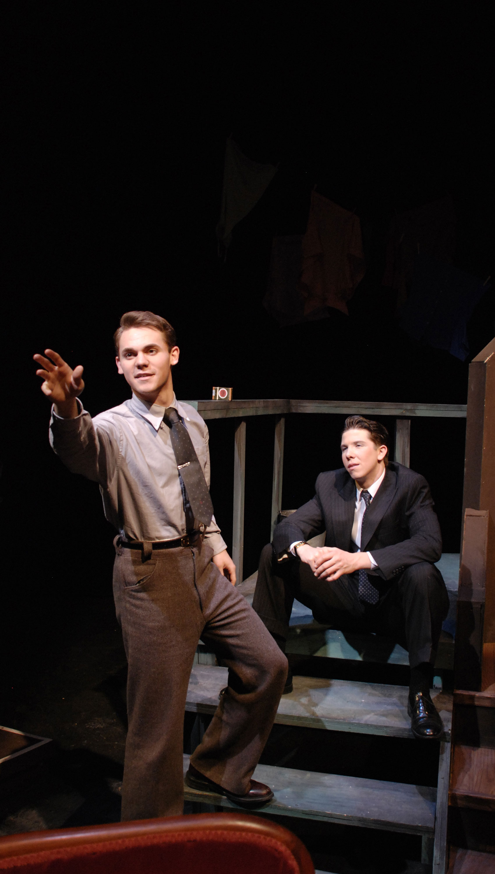 A young man stands, one foot on a porch step as he points out toward the audience, which he is facing, with his hand outstretched while another young man sits on a doorstep watching the first young man.