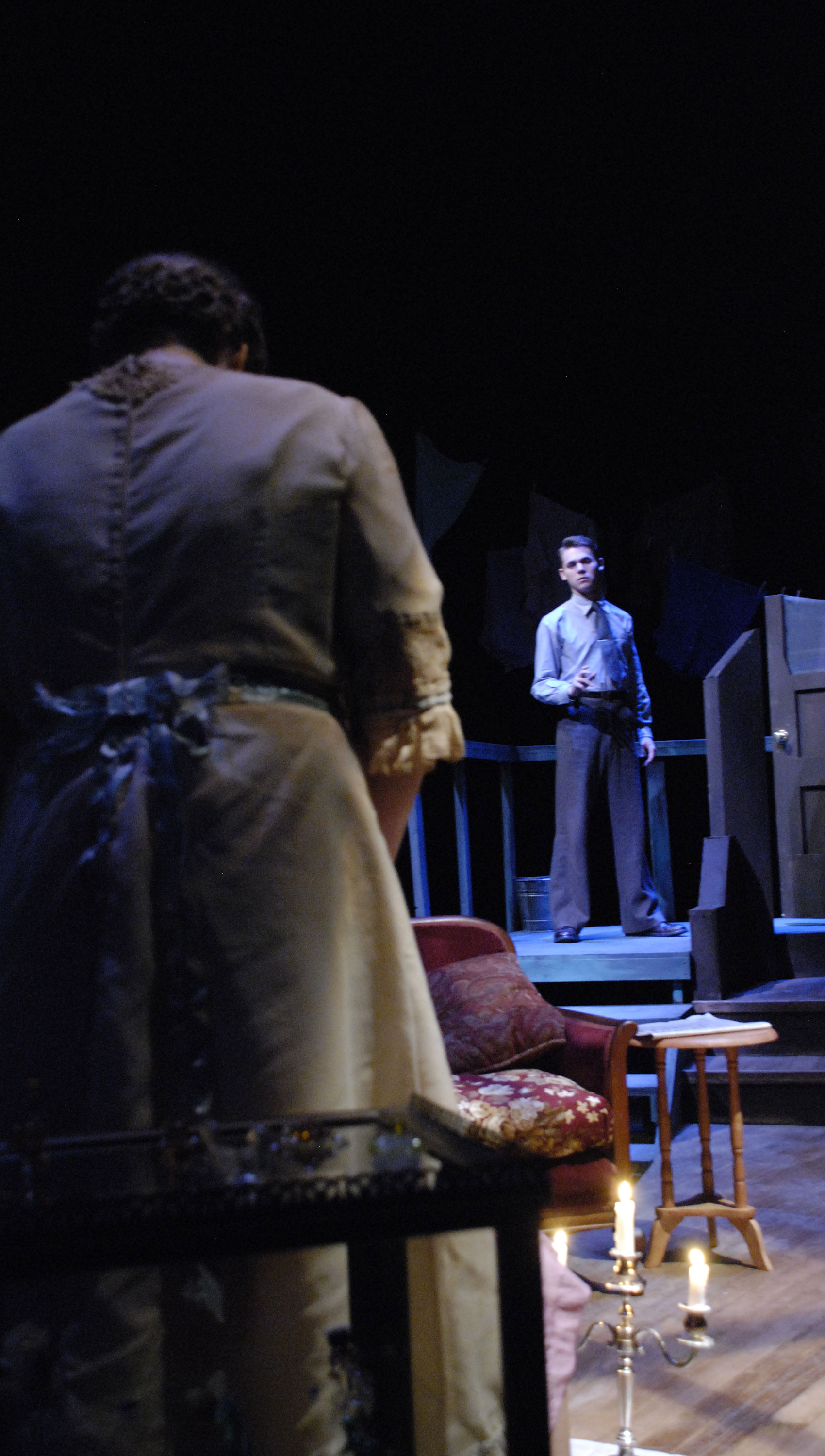 A young man stands on a doorstep further back onstage with his hand out low while a woman faces away from the audience further upstage, with her head bowed at the young man and her arms in front of her.