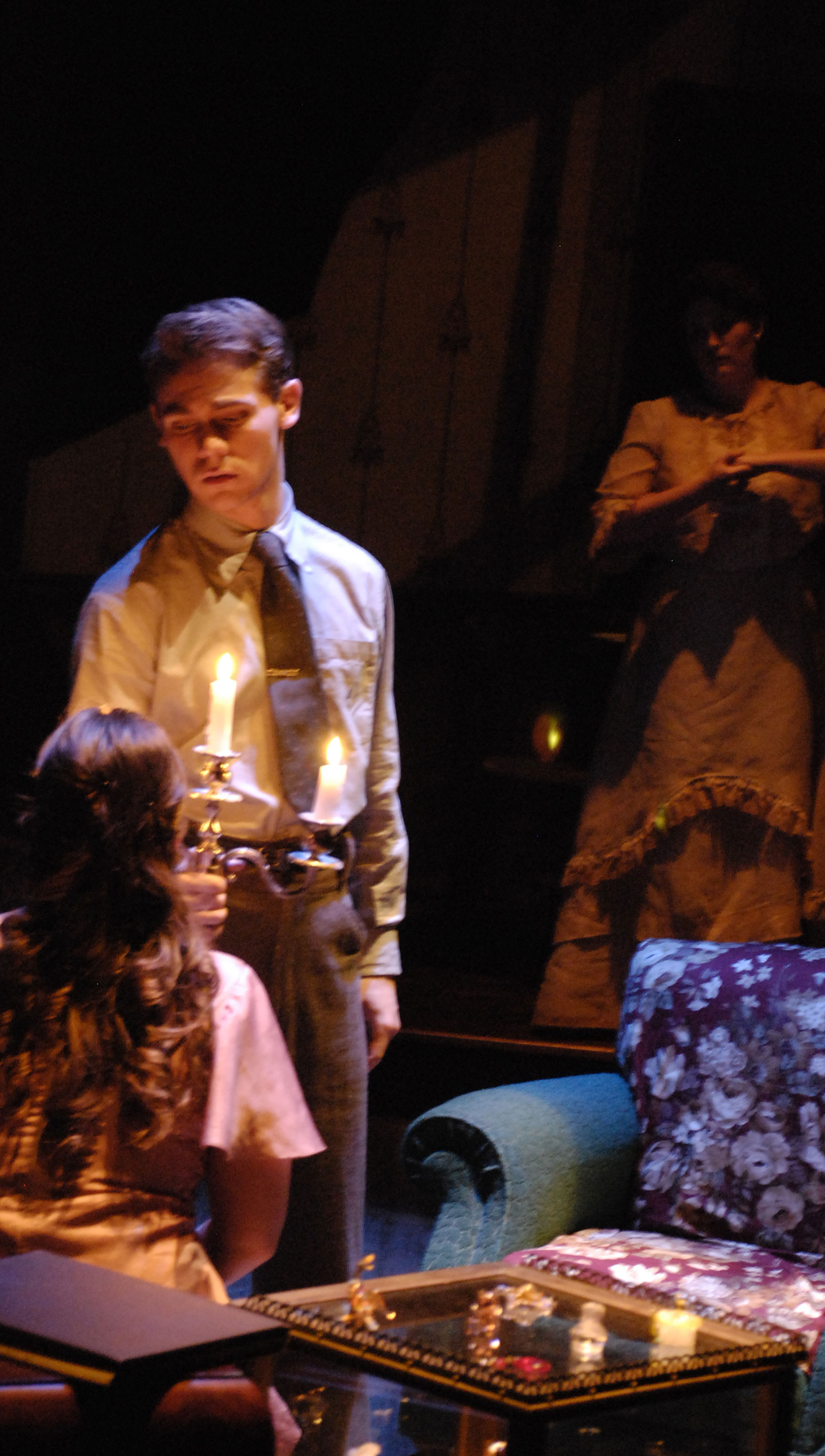 A young man stands in front of a young woman, who sits looking up at him, with a lit candlestick that illuminates them both, while an older woman watches the two from the shadows.