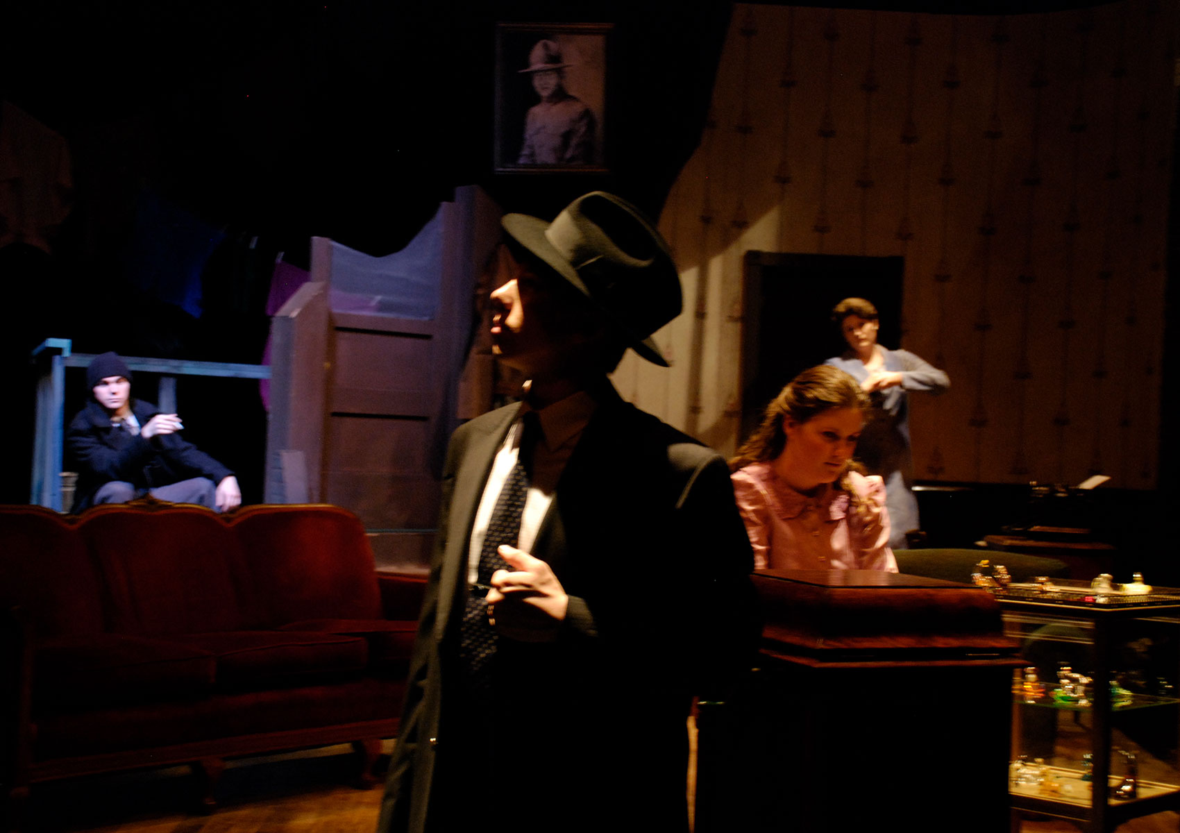 A man in a suit stands with his face in the shadows. Another man sits on the doorstep while an older woman stands behind a younger woman sitting in the sitting room.