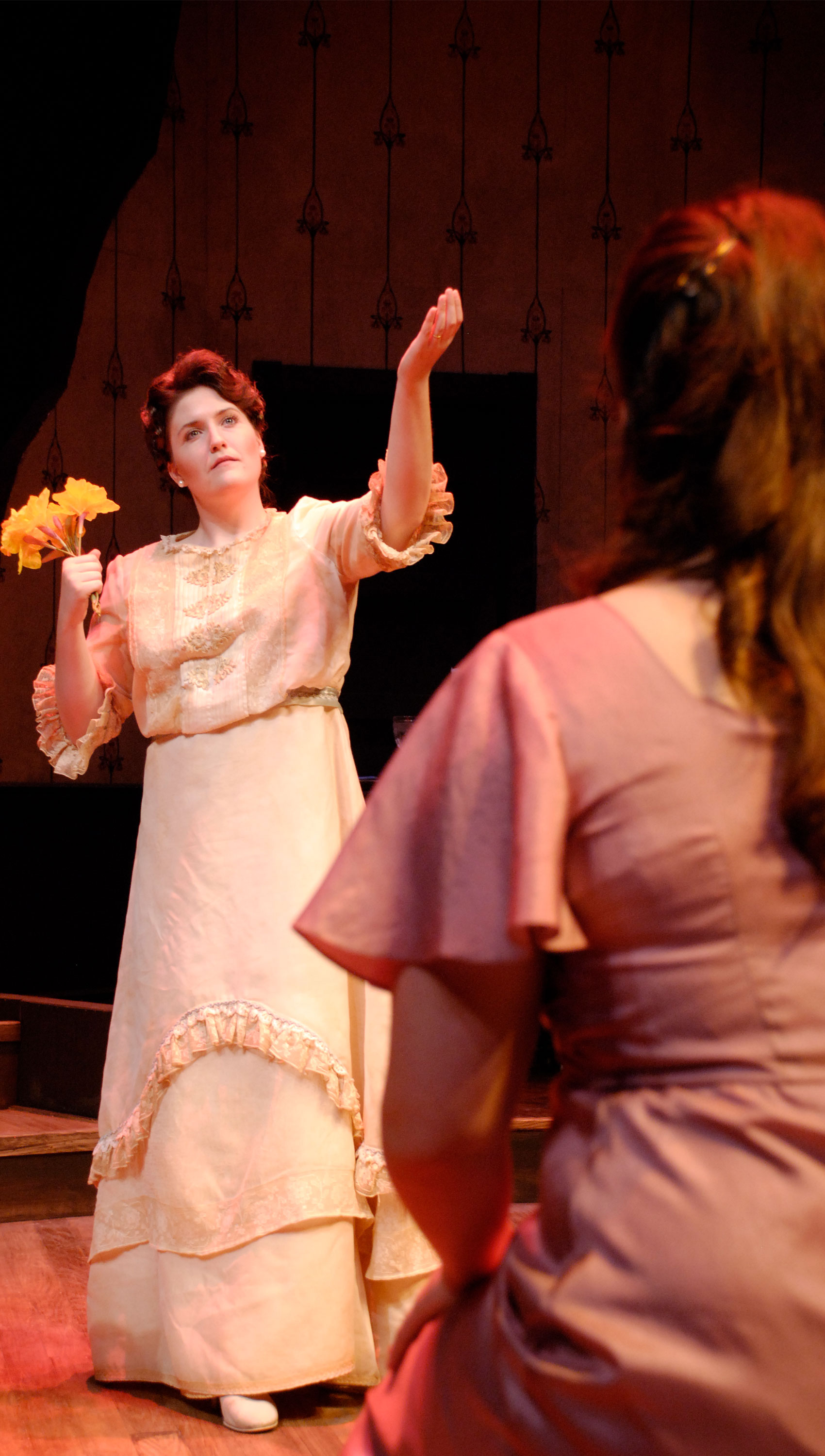 A young woman watches an older woman in front of her with a bouquet of flowers in one hand and her other hand stretched toward the sky; she is looking blankly over the young woman’s shoulder.