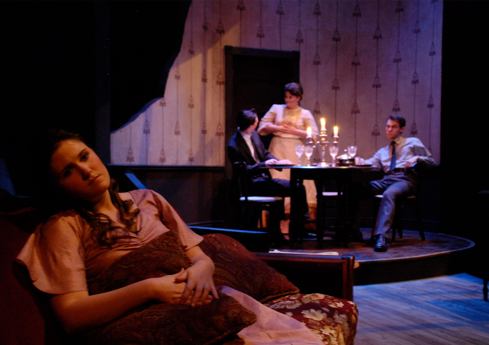 Two young men sit at a dining table while an older woman talks to  one of the two young men while the other looks annoyed A young woman sits on a couch further upstage while looking toward the audience, away from the three others.