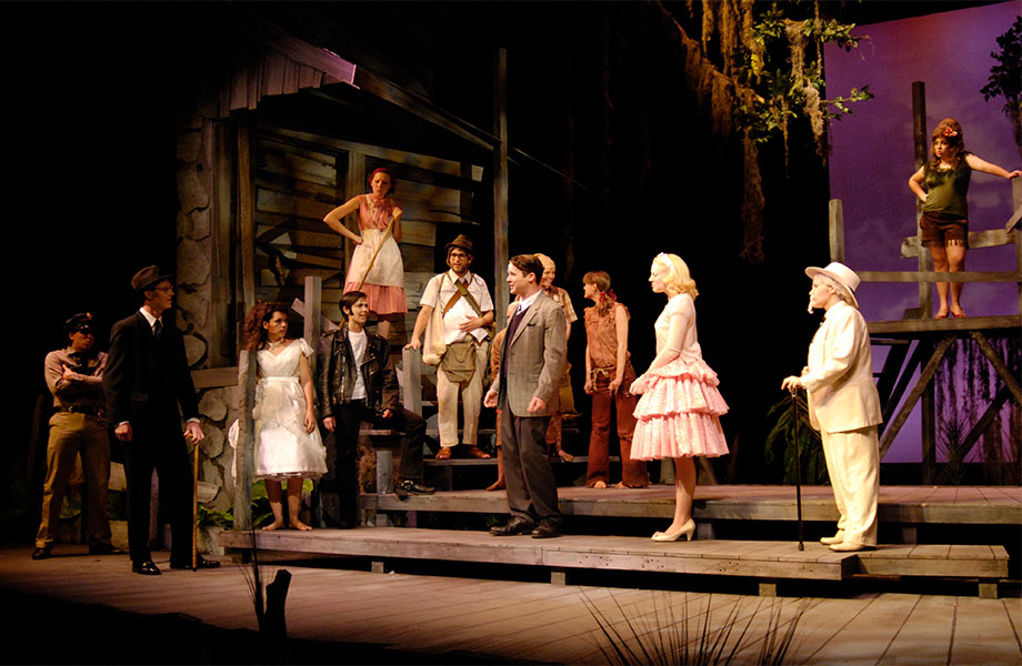 A group of differently dressed characters watch a conversation between two men taking place on the front porch of an old shack.