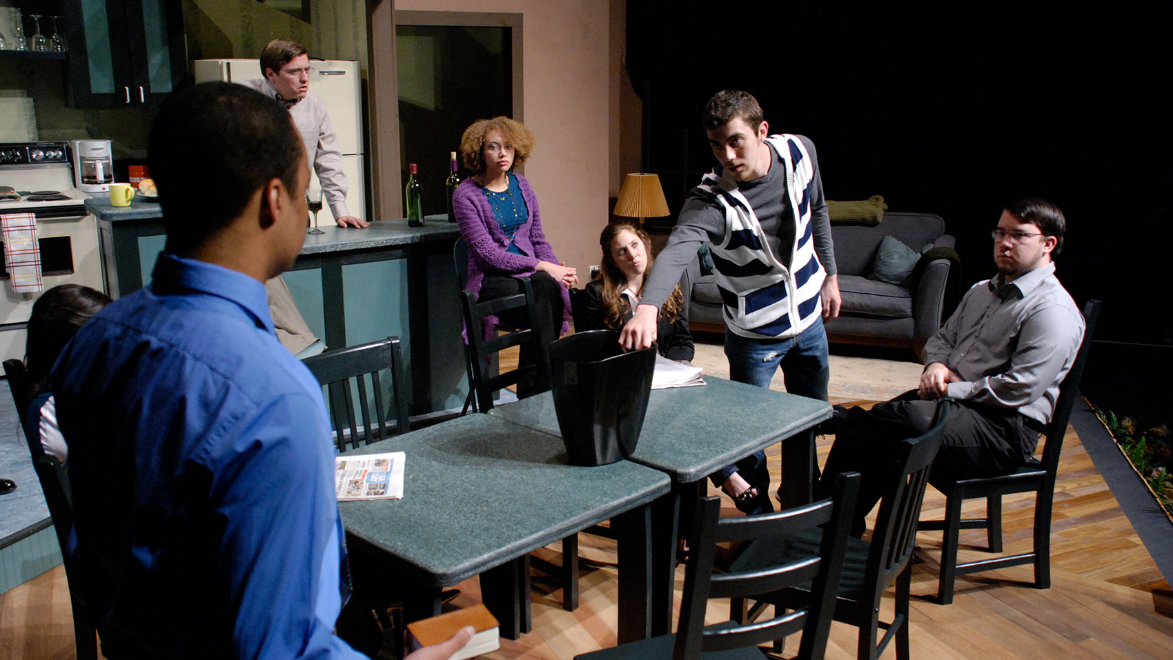A group of characters, some sitting and some standing, are surrounding a dining table. A man is standing gripping an empty black, plastic bin in the center of the table. 