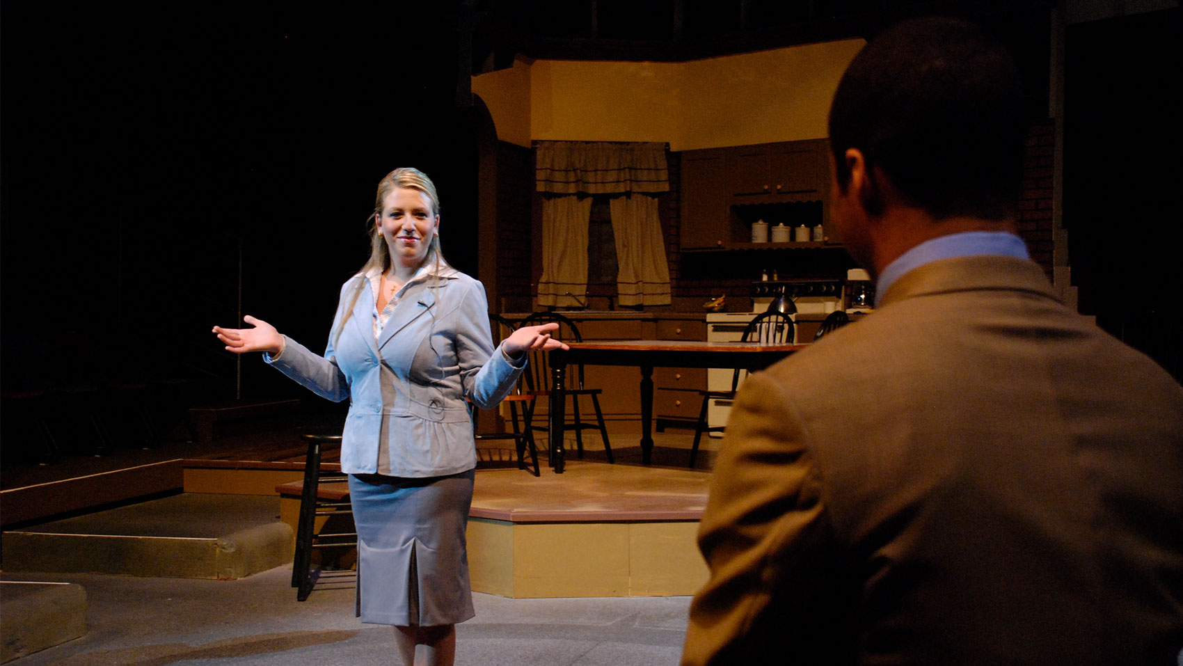 A man watches a woman, who stands in front of him with her hands up in a shrug, smiling. 