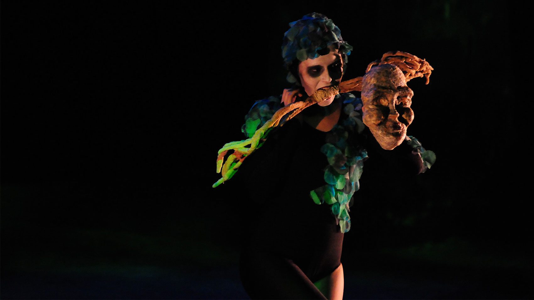 Someone in white face paint holding a large stick in their mouth and a mask