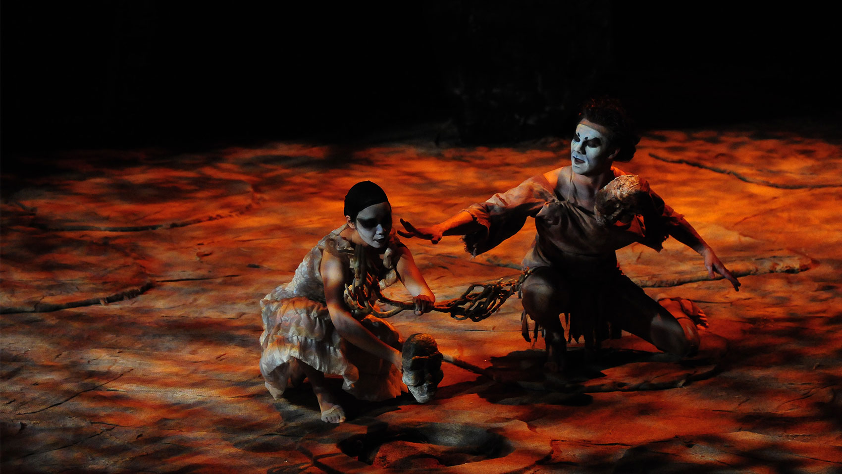 Two people in white face paint looking at a mask