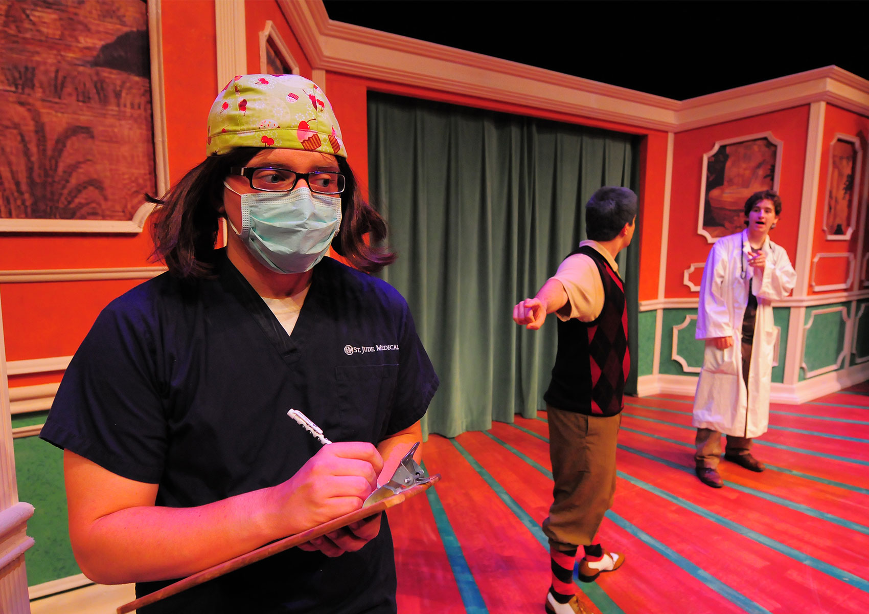 A doctor is talking to a man as the man looks at the doctor but points at the nurse upstage closer to stage right further behind him. 