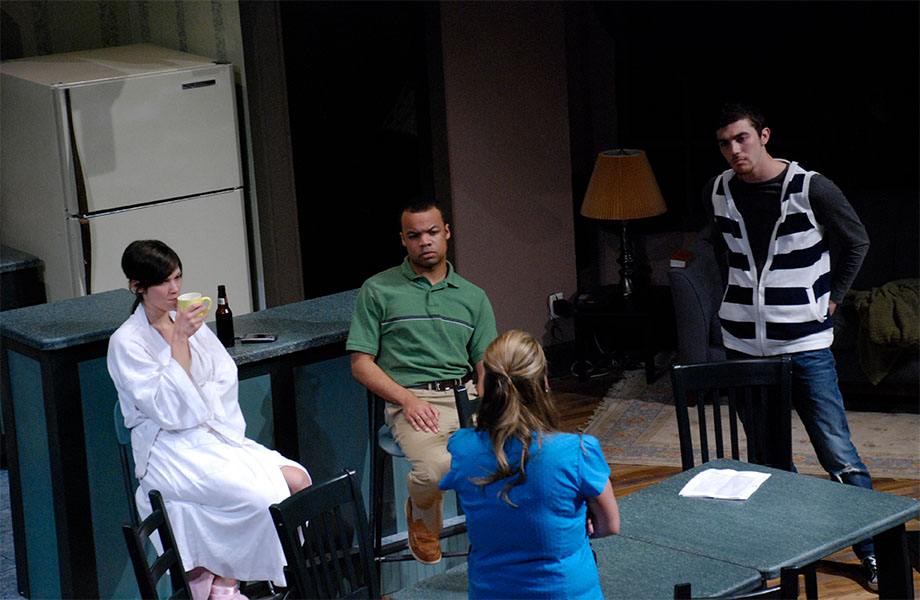 Three characters watch a fourth while she is speaking while they are all in a kitchen connected to a living room.