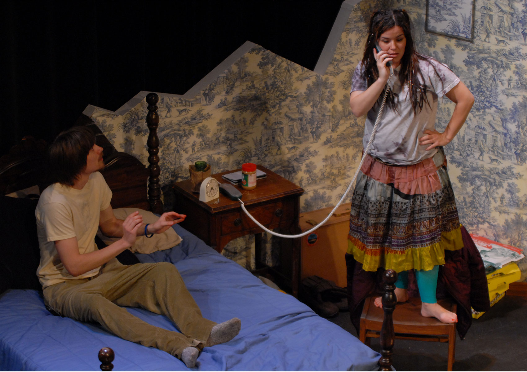 A man sits on a bed with his hands held out, a cheese curl in his right hand, while watching a woman standing next to the bed on a chair while talking on a landline phone.