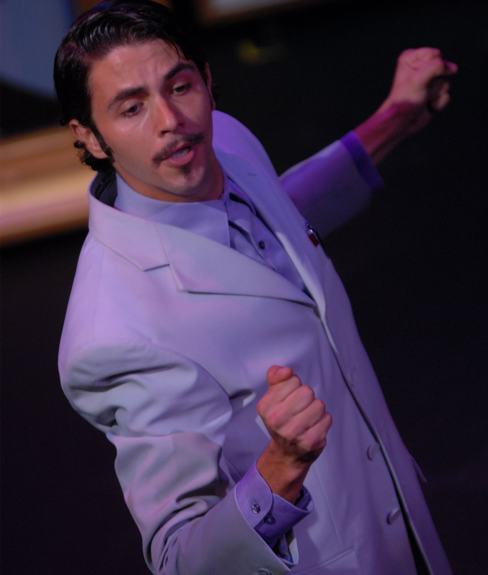 A man stands as if dancing while facing the audience, his chest outward toward stage left with his right hand in a fist in front of him and his left behind him.