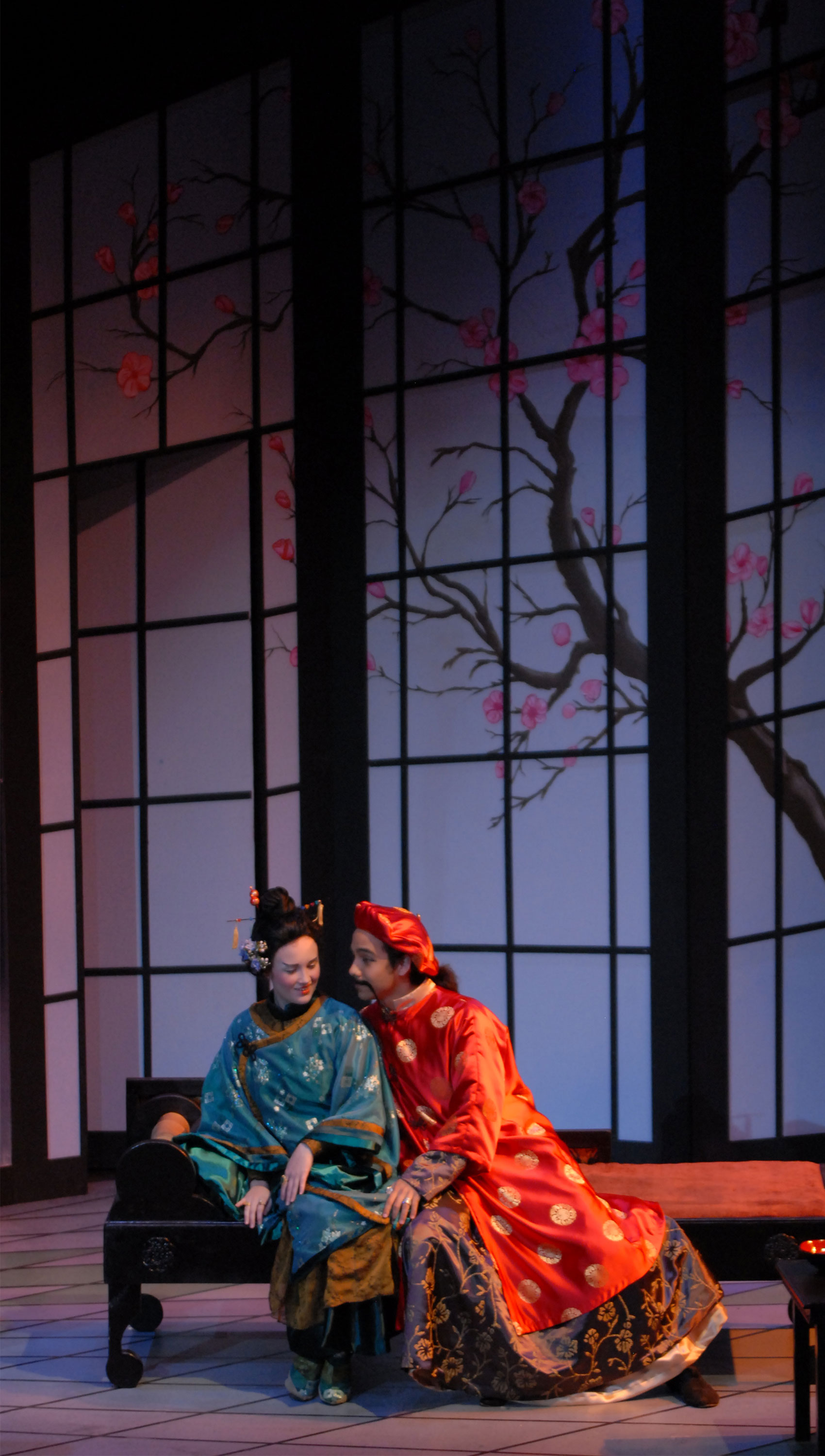 A woman dressed in Asian-inspired fashion and heavy makeup is on a bench next to a man, she is looking downstage coyly as she leans slightly away from him. The man is also wearing fashion reflective of Asian culture and is staring at the woman while smiling.