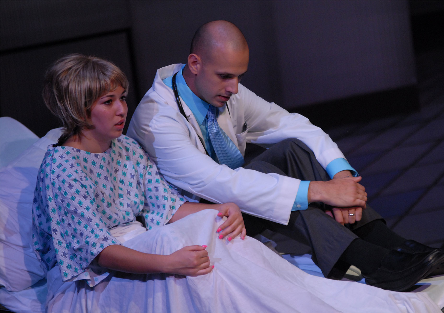 A woman sits in a hospital bed covered in a cloth while holding her knees, staring outward. A man dressed as a doctor sits next to the woman, looking down.