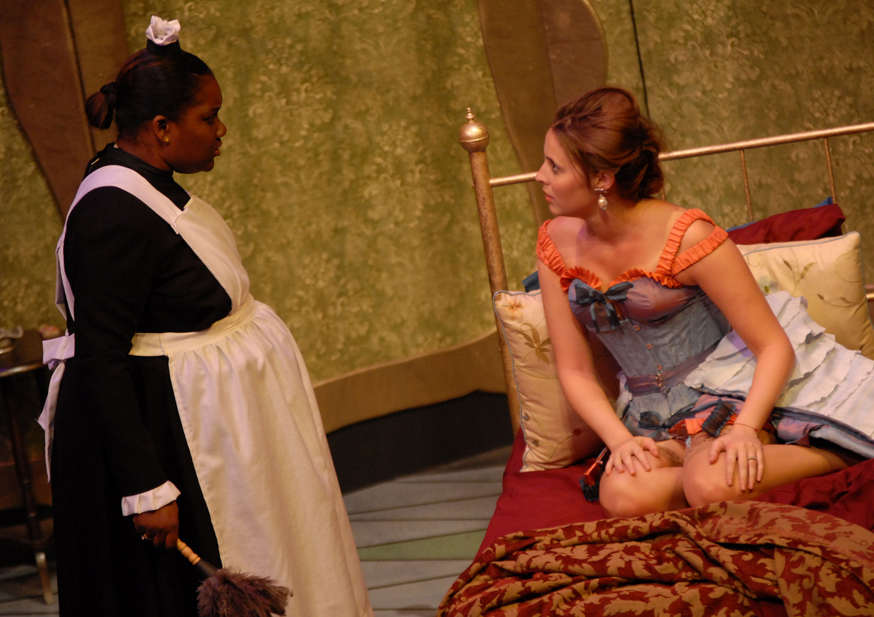 A woman dressed as a maid is standing and in conversation with a woman dressed in lingerie, who is sitting on her knees in a bed that is unmade; this woman is dressed in lingerie.