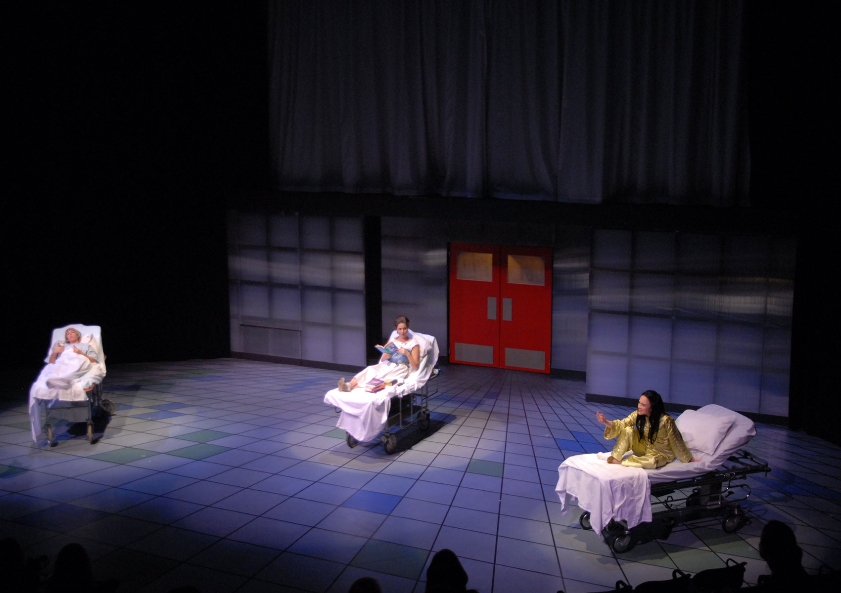 Three hospital beds are spread far apart on stage with a woman occupying each. The woman in the bed farthest left is laying, looking toward the audience. The woman in the middle bed is sitting up, reading. The woman in the bed farthest right is sitting up and gesturing.