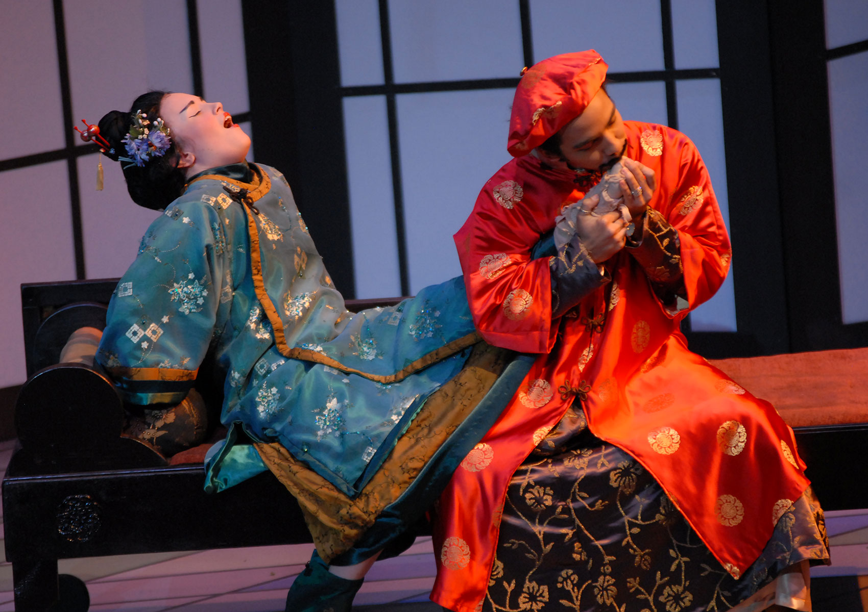 A man in Asian-inspired attire is sitting on a bench, kissing the bound foot of a woman next to him dressed in Asian-inspired attire; she is leaning back with her mouth open and her eyes closed.
