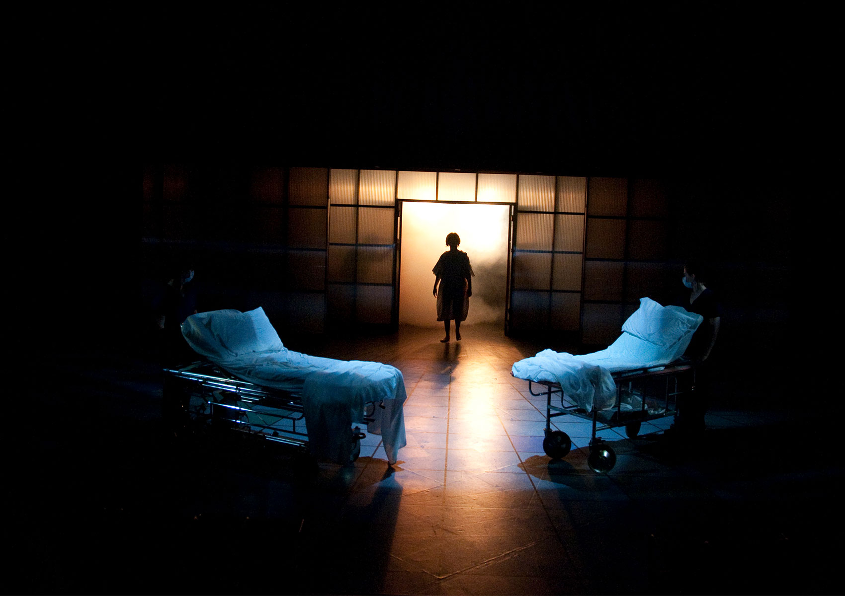 Two unmade hospital beds are toward the middle of the stage. A character walks, silhouetted, toward an open door filled with golden light.