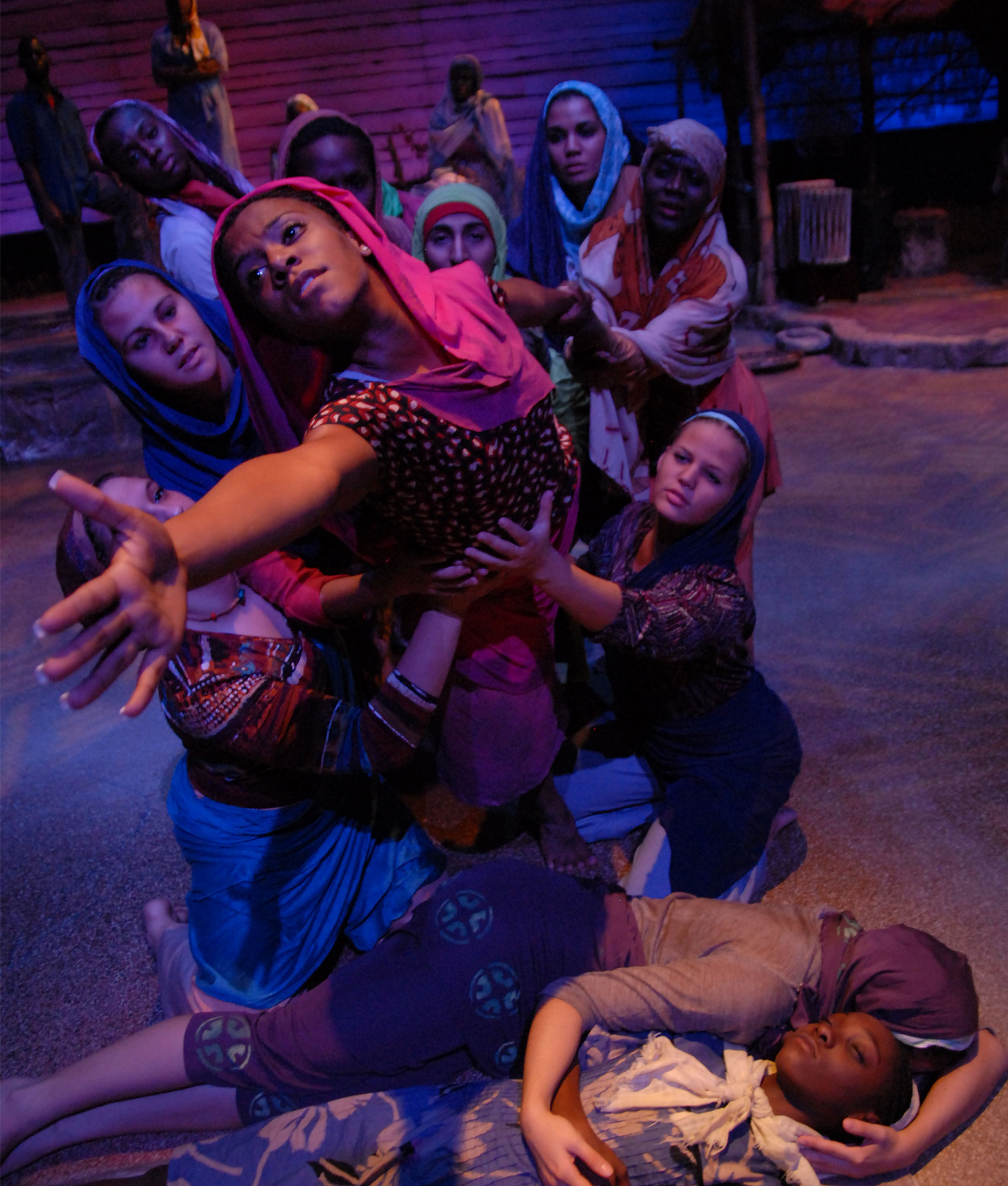 A child lies on the floor, embraced by someone, while a group of people surround another woman