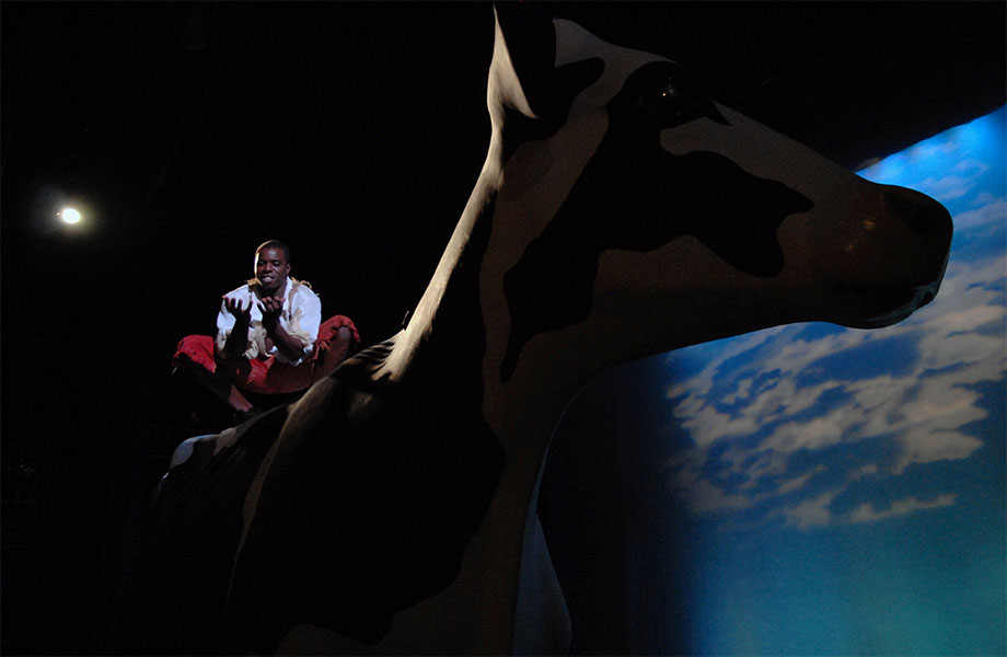 A man has his palms outstretched while squatting on the back of a giant cow in front of a backdrop of the sky.
