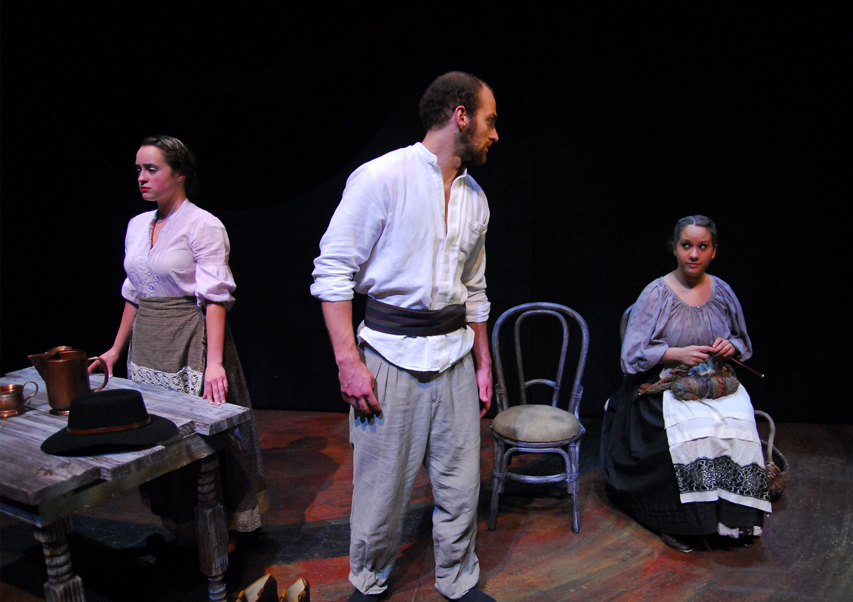 A man stands with two women spaced farther apart on either side of him. The woman to the man’s left is looking forlornly downstage while placing her hands gently on a wooden tabletop that has a cup, pitcher, and hat on top of it. The man is looking right toward the woman on his right side and the woman, who is sitting in a chair next to an empty chair while knitting, looks sideways back at the man. 