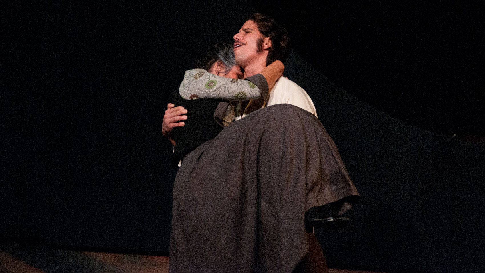 A man carries a woman in his arms. Her arms ae wrapped around his neck and her face is buried in his neck and he has his eyes closed and mouth ajar, as if he were exclaiming or had been crying.