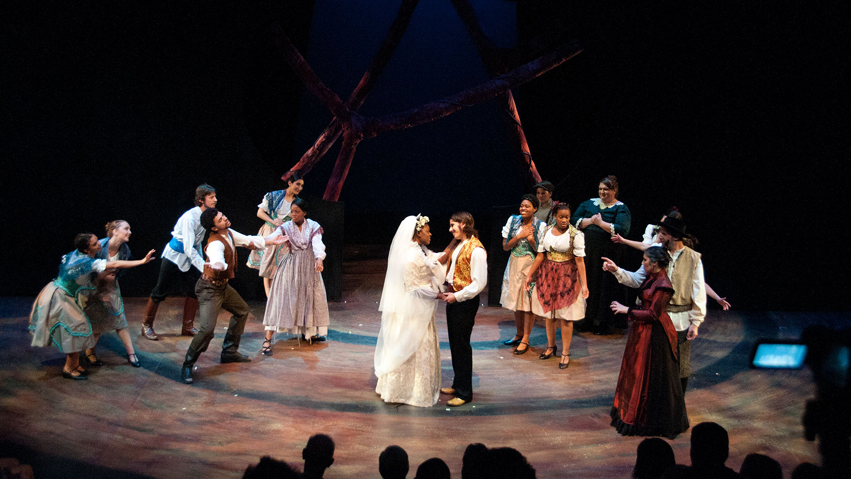 A bride and groom hold each other, staring into each other’s eyes lovingly as onlookers look on happily, some gesturing at them and some just standing by. 