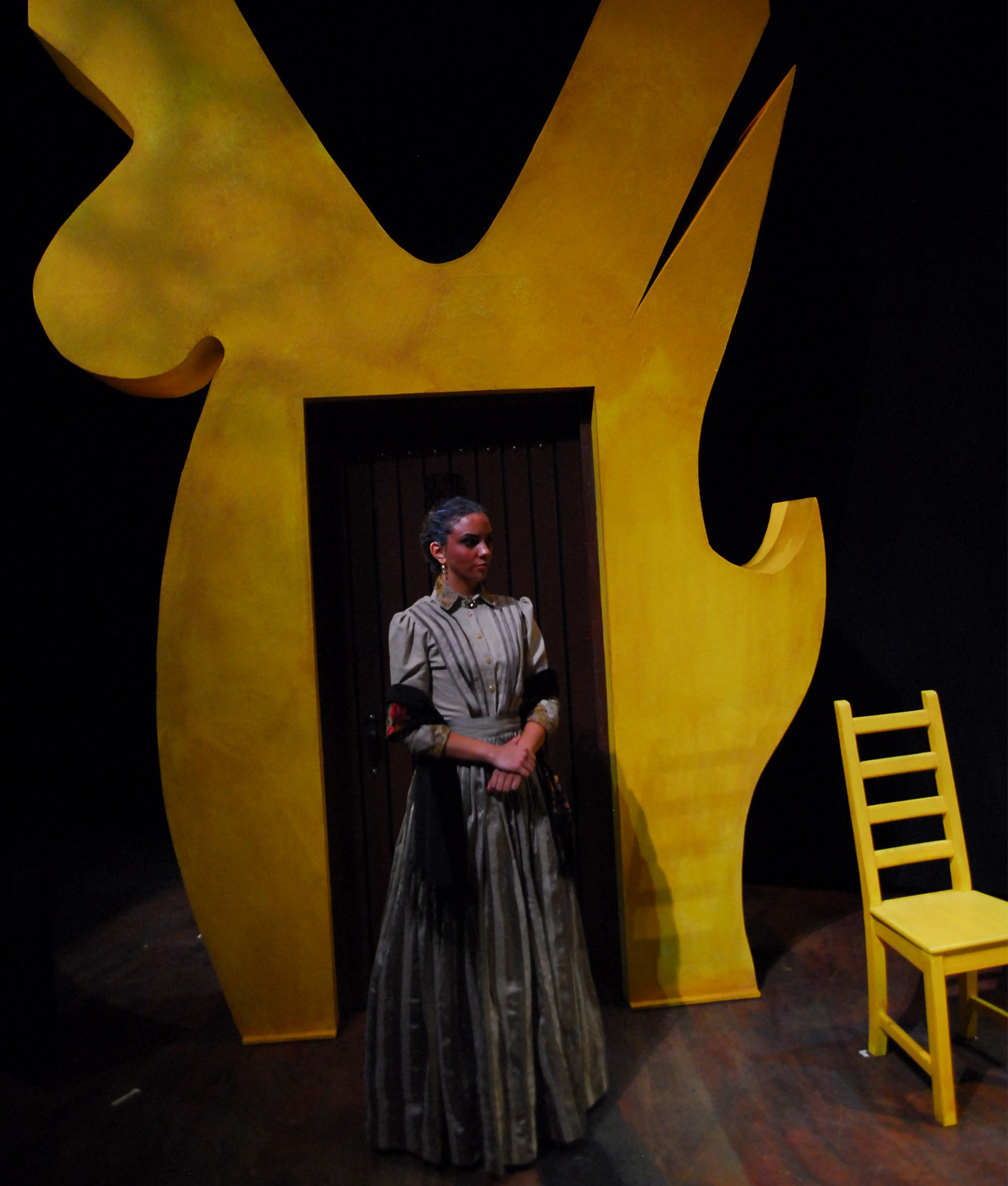 A woman stands in front of an abstract-shaped yellow doorway, which is positioned next to a yellow chair. 