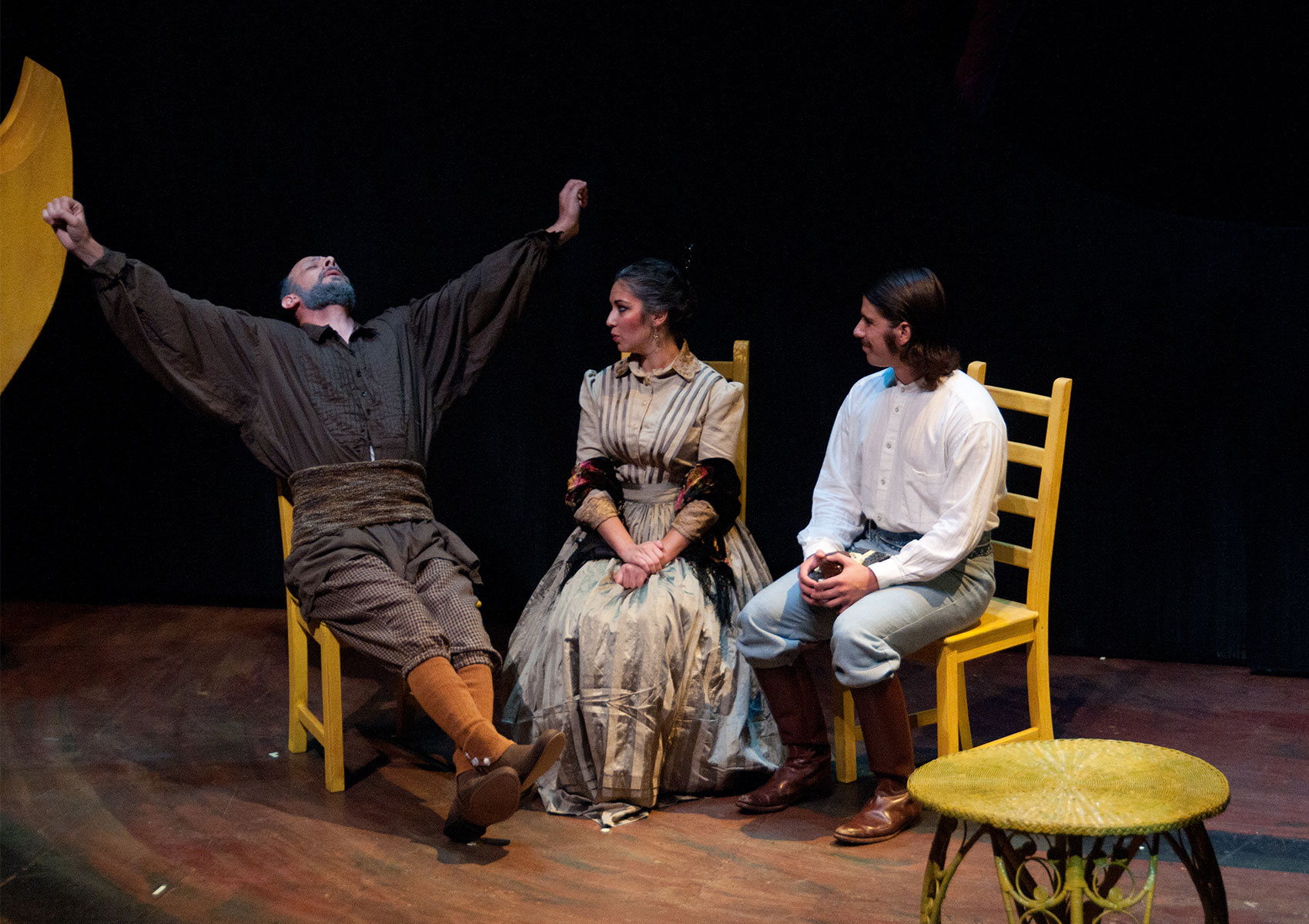 An older man sits next to a woman and man in left to right order, all three sitting in yellow chairs. The older man is stretching his arms out, crossing his legs, and leaning back as if he were tired. The woman and man are watching him. 