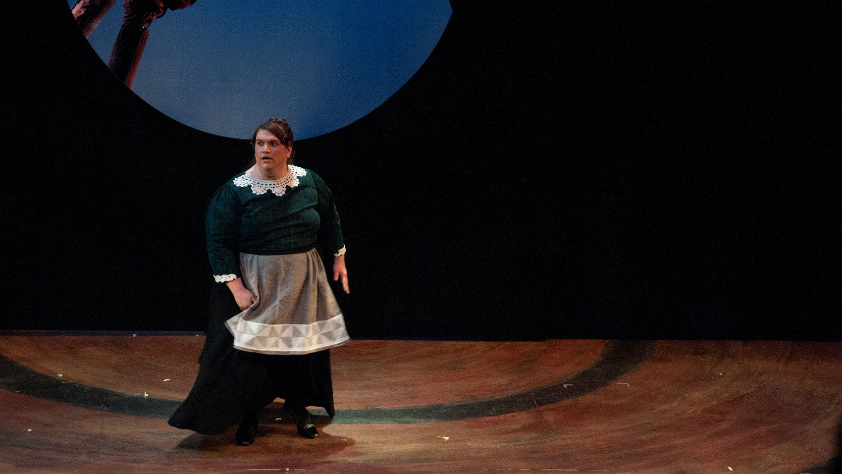 A woman dressed as a maid stands alone on stage.