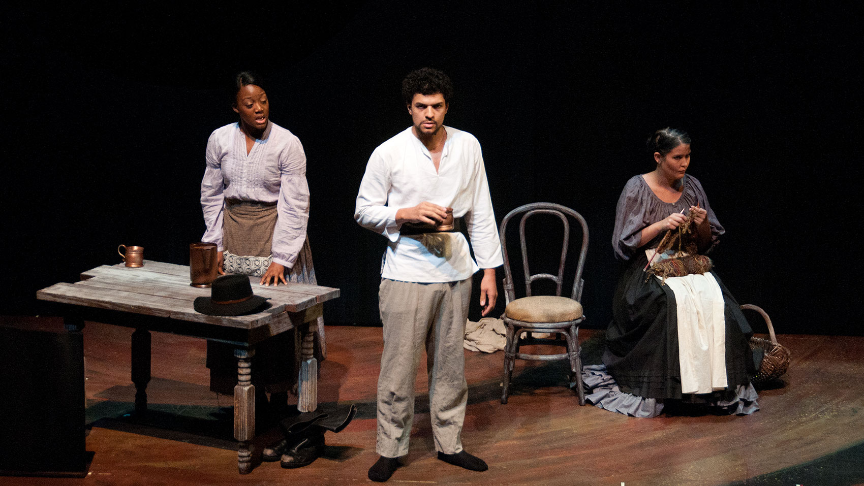 A man stands in between two women spaced apart on either side of him. The woman to the left of the man is speaking while placing her hands gently on a wooden tabletop that has a cup, pitcher, and hat on top of it. The man stares intensely toward the audience holding a cup while the other woman sits in a chair next to an empty chair while knitting.