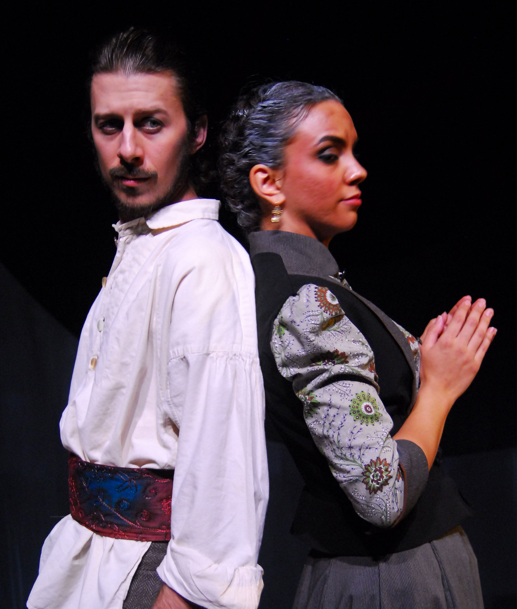 A man in a white shirt and vibrant red and blue sash stares outward on an oblique angle toward the audience. He has his back pressed against a woman’s, who is wearing a gray and black dress with her palms pressed together as she leers to the side toward the audience. 