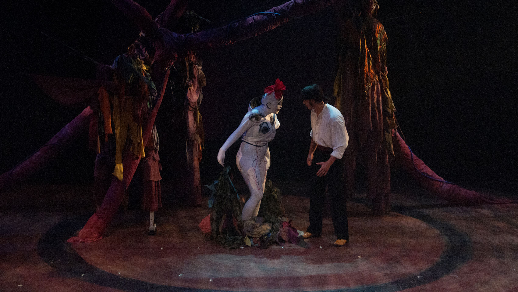 A man and a creature are having a confrontation under an arbor of trees. The man is wearing a white shirt with the sleeves rolled up and black pants as he is confronted by a creature that is solid white with a red flower on its head and has three symbols resembling eyes drawn onto the area near its chest. It is holding blankets that are green and brown textured, resembling foliage.
