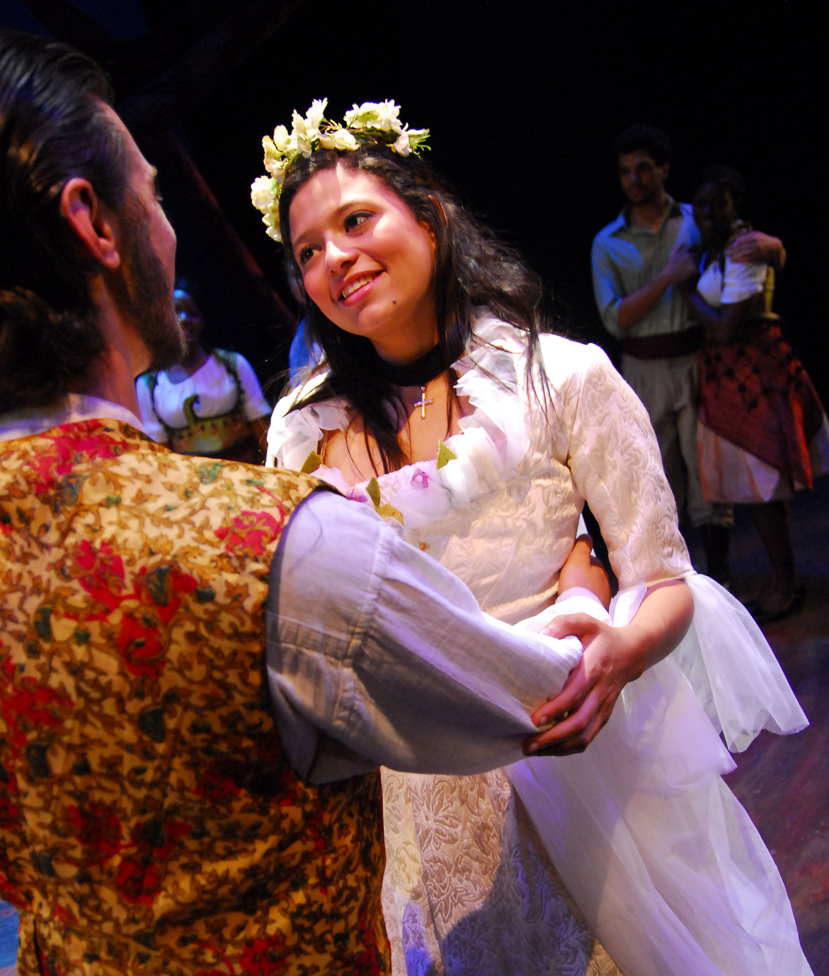 A bride is held by her groom and is looking up at him lovingly, a couple holds each other and watches them happily, slightly out of focus in the background.