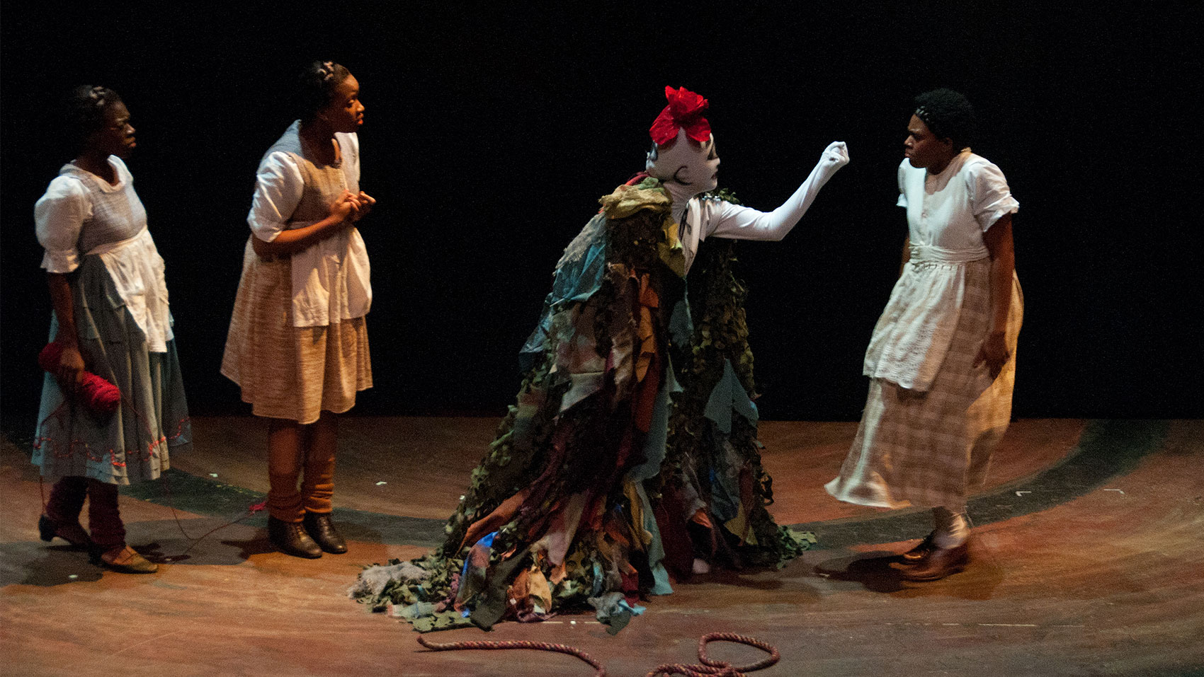A creature that is solid white, covered in drawn symbols and a red flower on top of its head is wearing a cloak that resembles foliage and is holding a closed fist toward a woman who looks visibly frightened. Two women stand behind the creature, one talking and gesturing downward with her index finger while the other holds an unspooled roll of red yarn and looks on with her brow furrowed as if confused. 