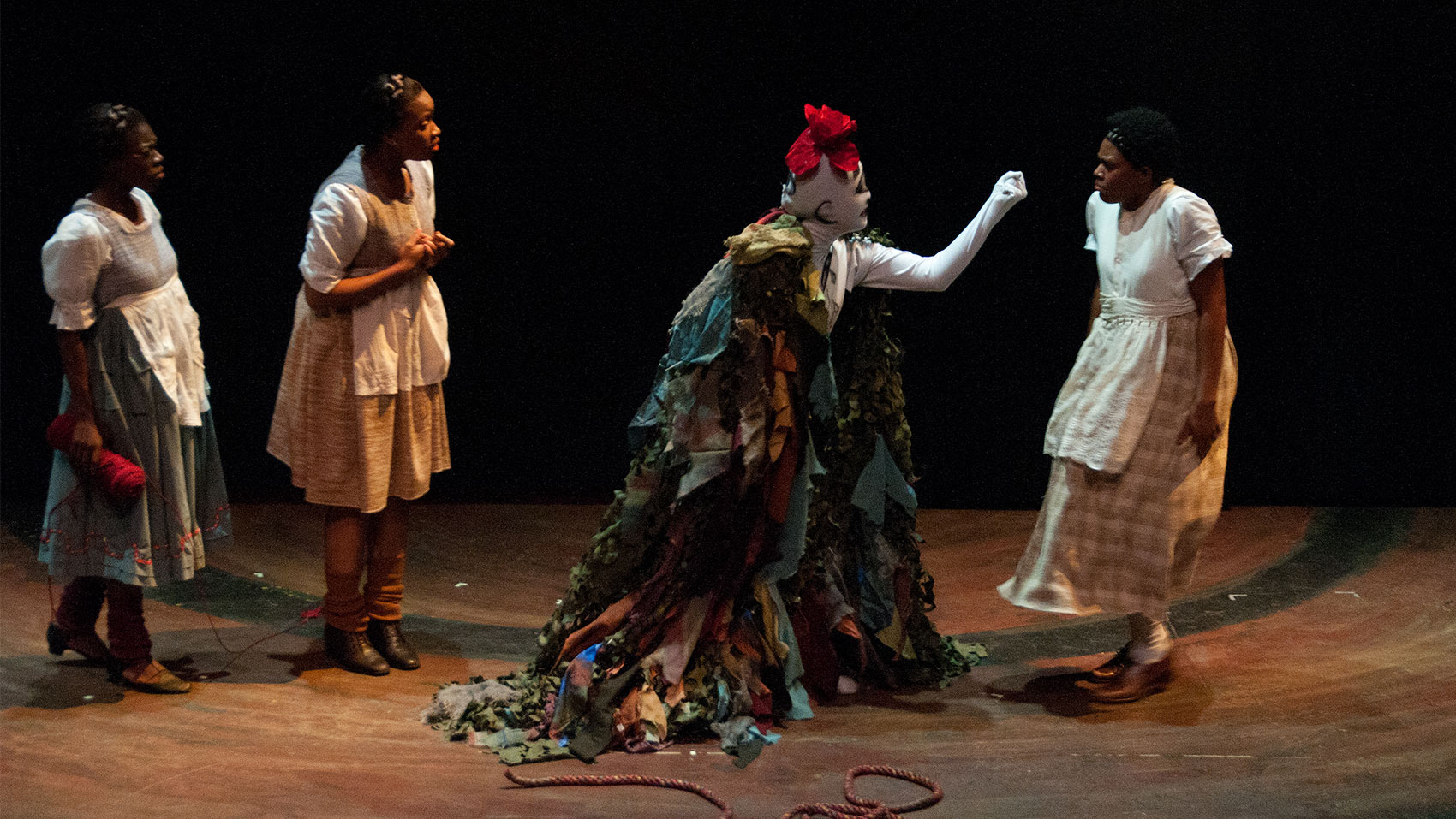 A creature that is solid white, covered in drawn symbols and a red flower on top of its head is wearing a cloak that resembles foliage and is holding a closed fist toward a woman who looks visibly frightened. Two women stand behind the creature, one talking and gesturing downward with her index finger while the other holds an unspooled roll of red yarn and looks on with her brow furrowed as if confused.