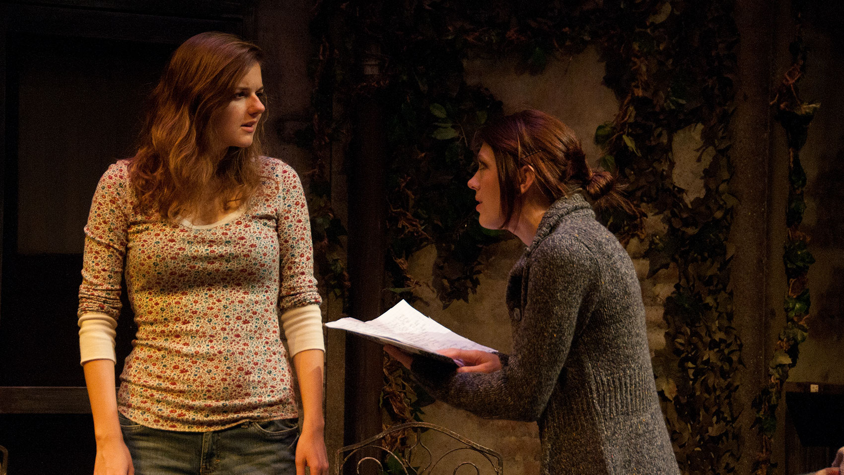 Two women are locked in conversation. One woman looks indignant as another woman is angling her neck toward the first as she holds open a notebook as if she were reading it aloud and paused to speak.